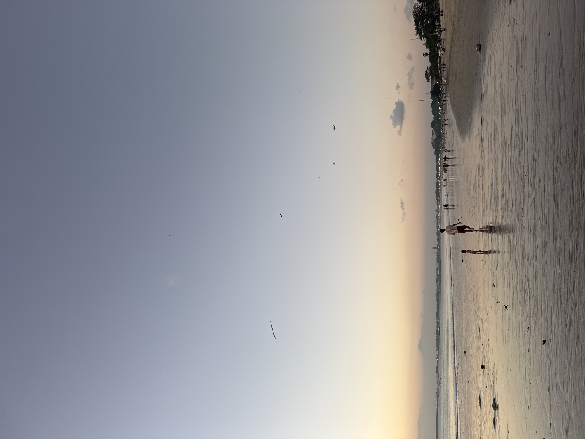 The image captures a wide shot of Kuta Beach in Kuta Selatan, Indonesia, during what appears to be sunset or dusk. The sky transitions from a light blue at the top to a warm yellow-orange hue near the horizon, indicating the late hour. Several small figures are scattered across the wet sand, which reflects the sky's colors. In the foreground, a person and a child are walking away from the camera towards the water, casting shadows on the sand. Further down the beach, a crowd of people are walking and gathering, with some sitting under umbrellas or awnings on the sand. The ocean is calm with gentle waves lapping the shore. In the distance, a line of buildings and trees forms the coastline, with a few lights beginning to illuminate. There are also several small dark shapes visible in the sky, possibly birds or kites.
