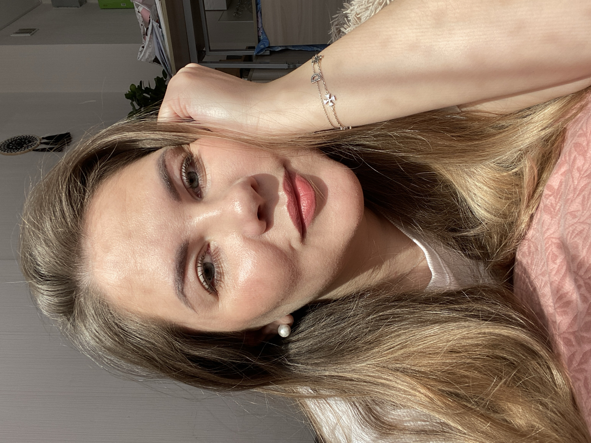 A light-skinned woman with long blonde and brown hair is positioned in the center of the frame. She is lying down, with her head resting on her left hand and arm, which is adorned with a thin, delicate bracelet. She is wearing a white top. The background is blurry and out of focus.
There is no indication that this image was taken in Izhevsk, Russia.