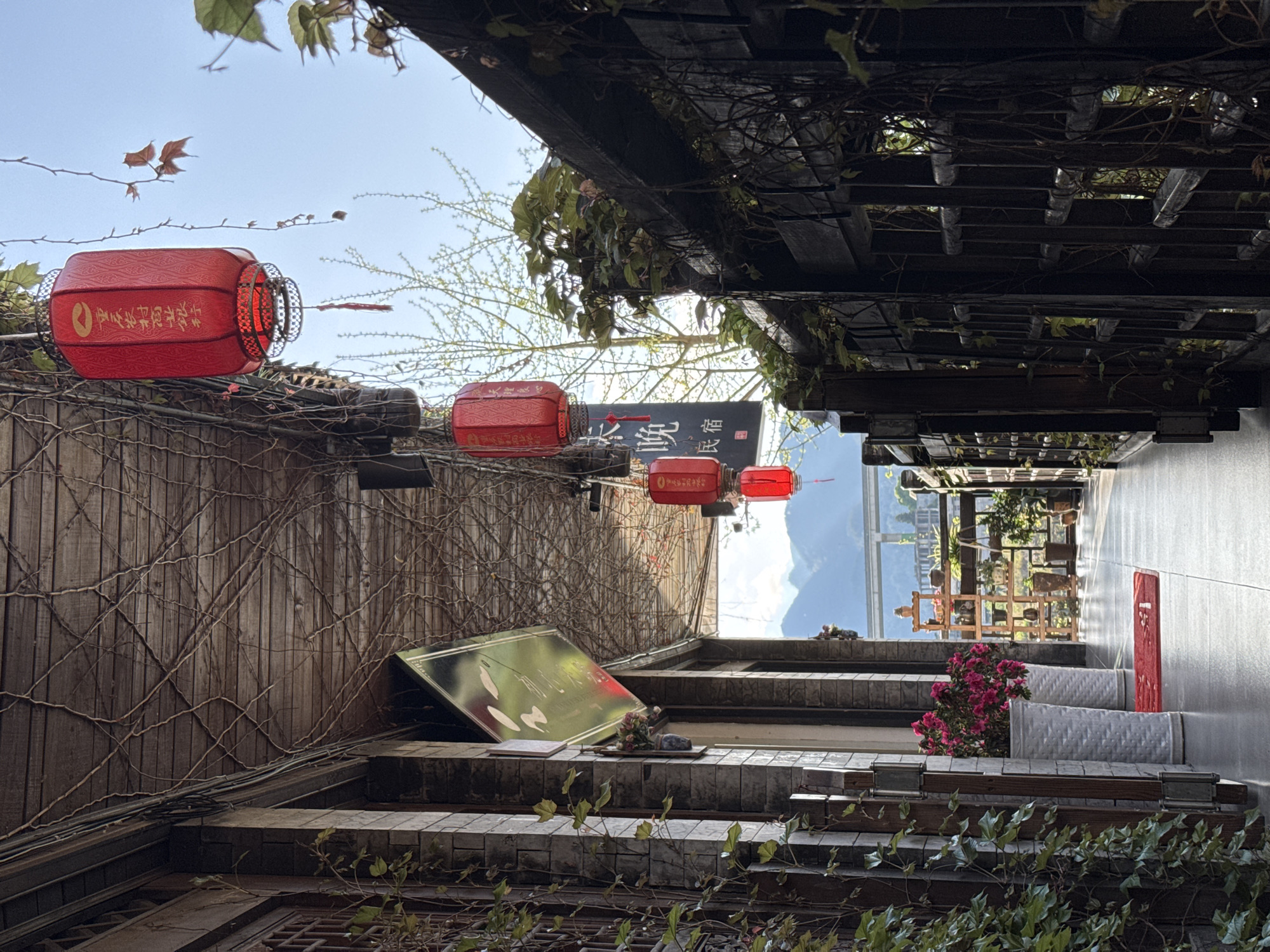 The image depicts a narrow alleyway in Zhuoshui, China. The perspective is looking upwards through the alley. Traditional red lanterns are suspended overhead, with Chinese characters visible on them. Buildings on either side of the alleyway are constructed of wood and brick, covered with vines. A wooden lattice structure forms a partial canopy overhead. A sign with Chinese characters is visible. In the distance, a glimpse of mountains can be seen through the alley. There is a wooden staircase. Flowers are in pots. A red object is visible next to the pots of flowers.