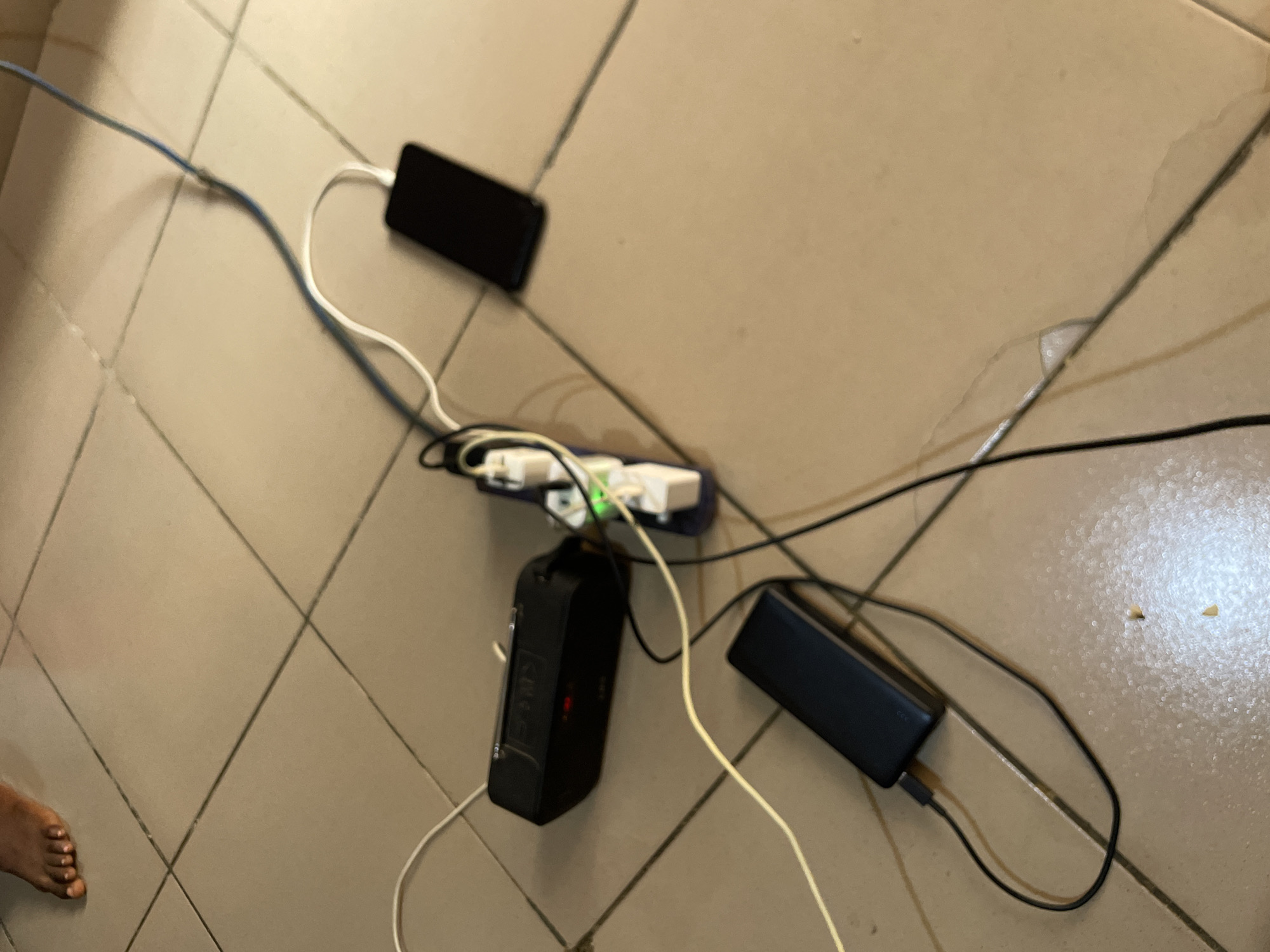 The image shows a tiled floor with several electronic devices plugged into a power strip. A black portable speaker is connected to the power strip, as is a black smartphone. A black power bank is also present, connected to a cable. A human foot with visible toes is in the upper left corner of the frame. The floor tiles are a light tan color and appear to be slightly wet in some areas. The scene is indoors, likely in a residential setting in Jos, Nigeria.