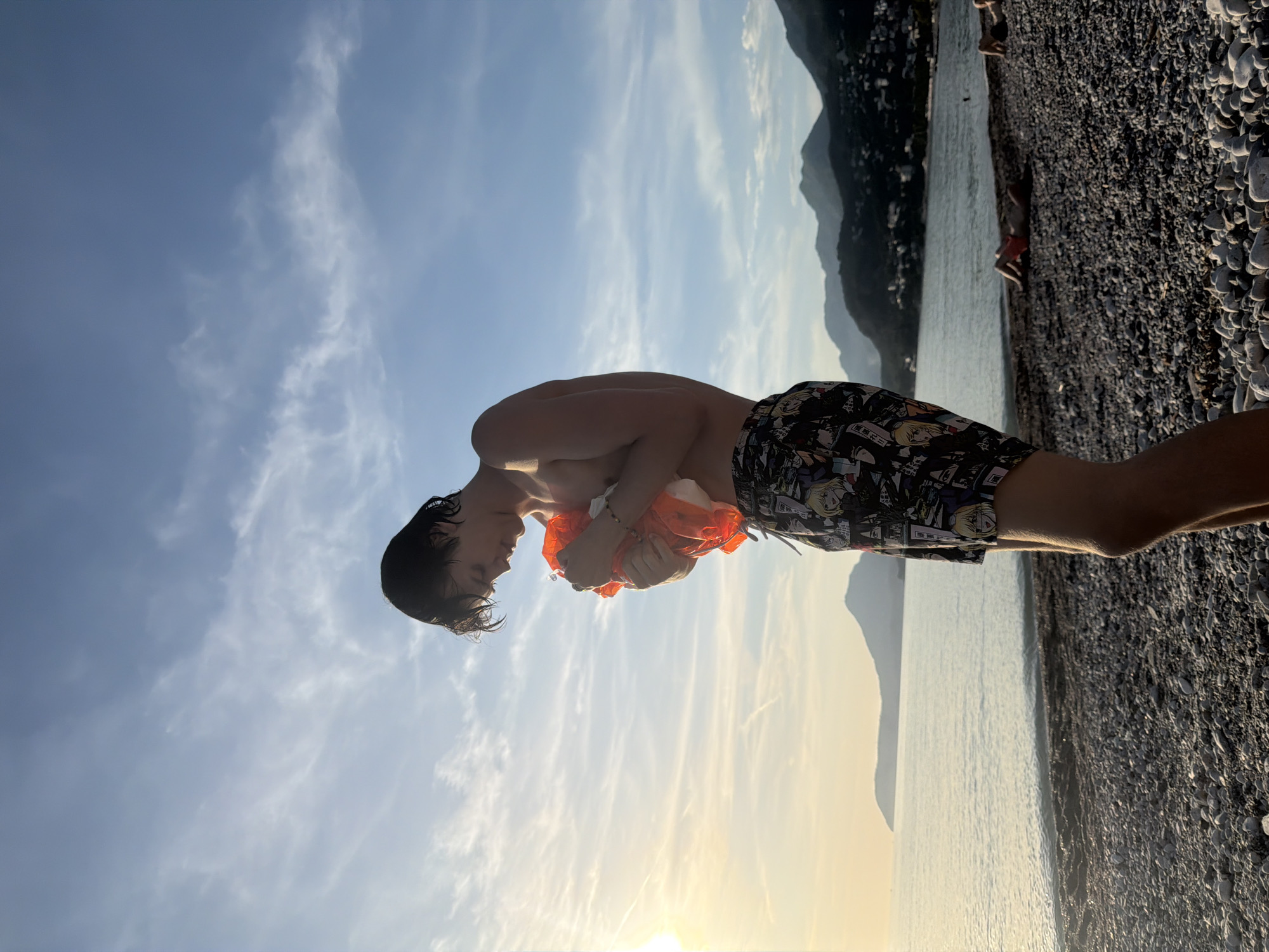 A young man stands on a pebble beach in Bar, Montenegro. He is bare-chested and wears patterned swim trunks. He holds an orange garment in his hands. Behind him, the Adriatic Sea meets the shoreline, with mountains visible in the background. The sky is blue with wispy clouds and the sun is setting. Other individuals are present on the beach in the background.