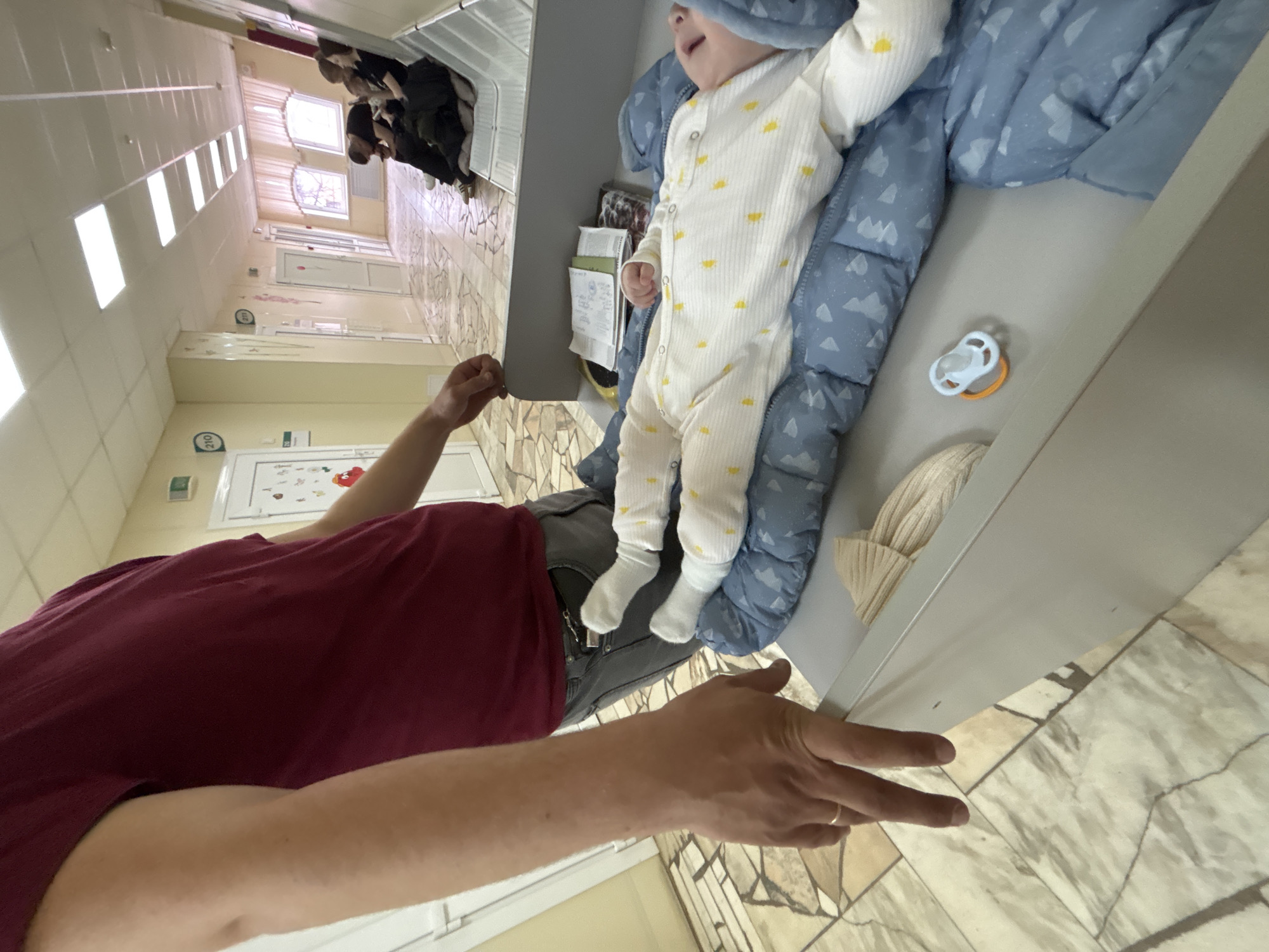 This image captures a low-angle, rotated view of an adult interacting with a baby within an indoor setting, likely a hospital or clinic.
In the foreground, an adult, dressed in a dark red t-shirt and grey pants, is seen from the chest down, reaching out with their left arm. A simple band-style ring is visible on their left ring finger. The perspective makes the adult's arm appear prominent.
To the right, a baby is lying on a blue blanket patterned with white clouds or mountains, placed on a grey surface resembling a crib, bassinet, or examination table. The baby wears a light-colored (white or cream) onesie adorned with small yellow sun patterns and white socks. The baby's face is partially visible, looking upwards, appearing to be vocalizing or making a face. A white and orange pacifier and a light-colored knitted hat are visible near the baby on the grey surface. Some papers, possibly medical notes, are tucked into a holder nearby.
The background reveals a long, brightly lit hallway with cream-colored walls and a tiled floor featuring irregular patterns of light and darker beige/grey. The ceiling has several rectangular fluorescent light panels. Multiple doors line the hallway, and through distant windows, natural daylight and leafless trees are visible, indicating it is daytime. In the far background, several individuals are present, appearing somewhat blurry, suggesting a waiting area or common space. Two signs displaying the number "210" are visible on the wall, likely indicating a room or ward number. A colorful poster with illustrations, including what appears to be a red apple or heart shape, is partially visible on the wall.
The scene suggests a moment of care or interaction between the adult and baby in a functional, well-lit public or institutional environment.