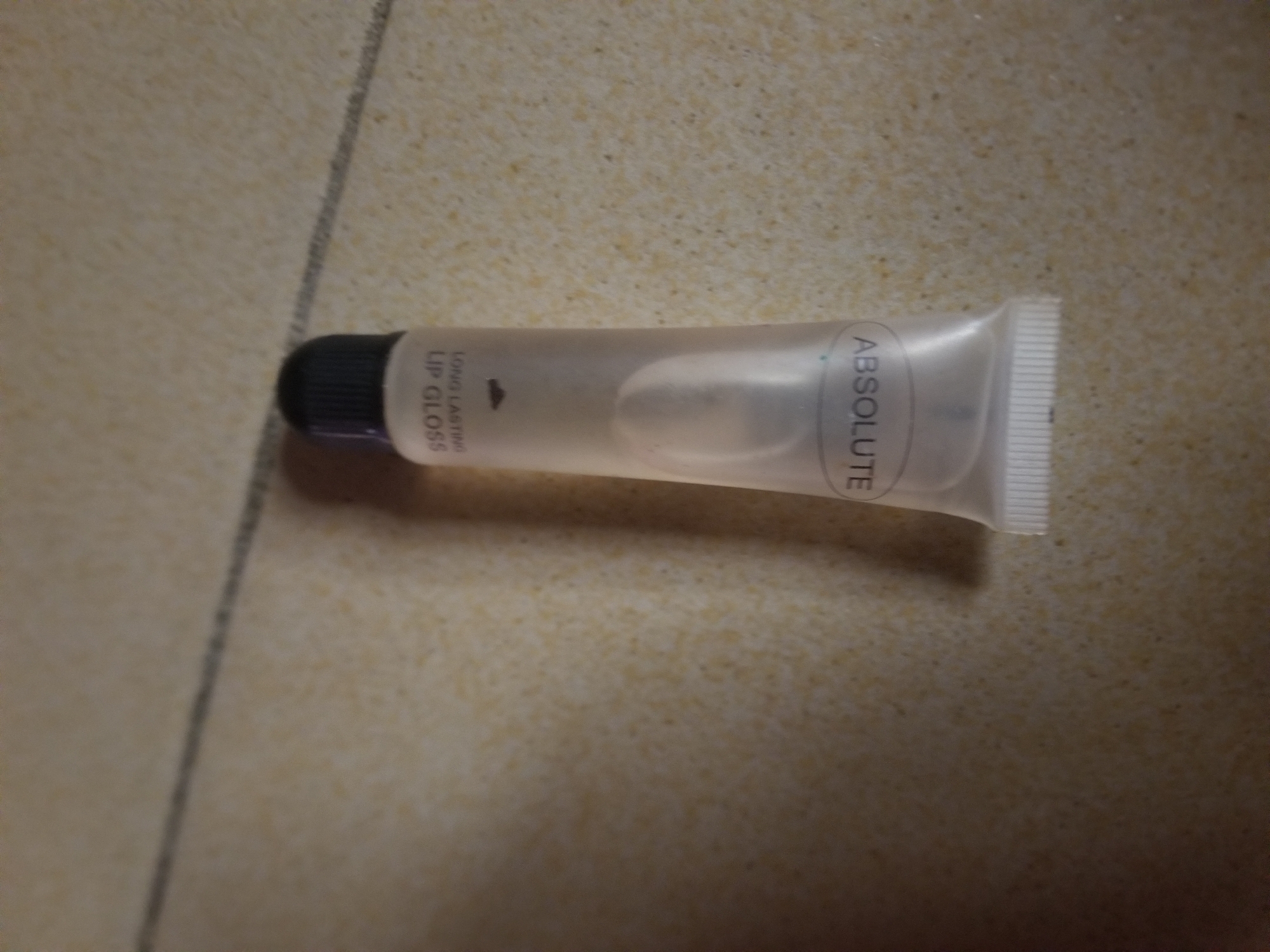 The image shows a tube of lip gloss lying on a tiled surface. The lip gloss tube is clear and has a black cap. Text visible on the tube reads "LONG LASTING LIP GLOSS" and "ABSOLUTE". The tiling appears to be beige or cream-colored with darker speckles. The lighting suggests an indoor setting. There are no people or animals present, and no specific activity is taking place. The image is a close-up shot of the object.