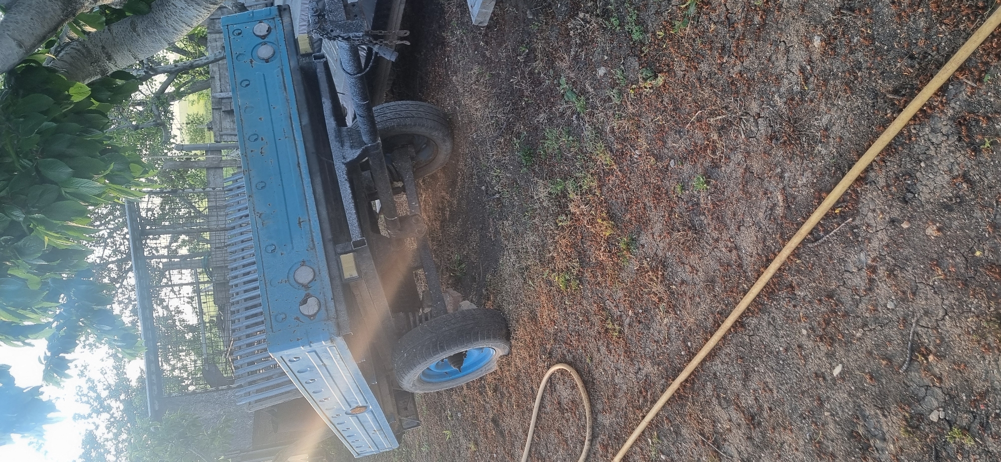 The image shows a blue flatbed truck in an outdoor environment in the city of Unknown, Unknown. The truck has a black undercarriage and blue wheels. A yellow hose lies on the ground near the truck. A tree with green leaves is visible in the background, as is a fence. The ground consists of dry soil with some vegetation.