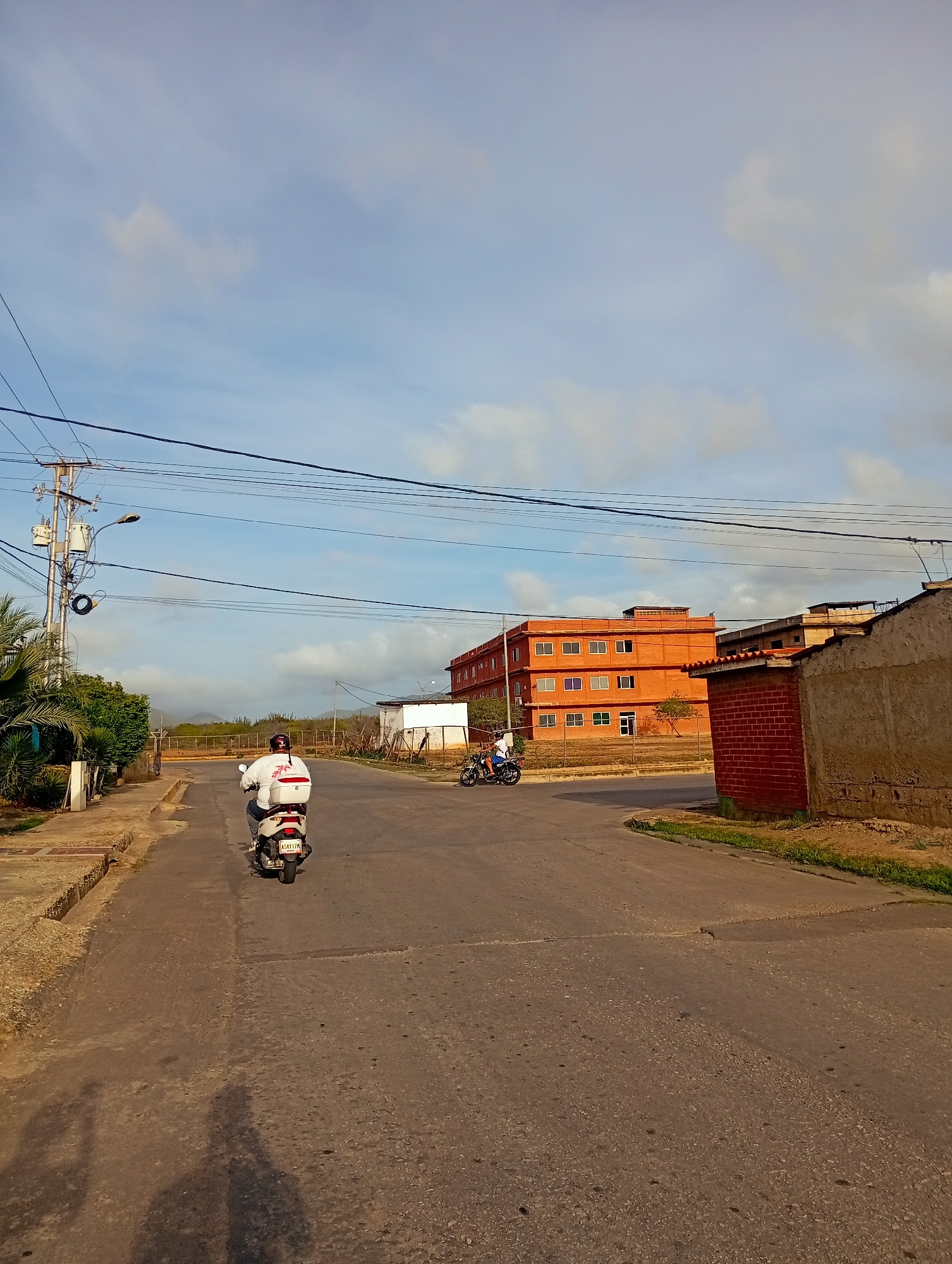 The image depicts a street scene in Juan Griego, Venezuela, with a clear sky and scattered clouds. A person on a white scooter, wearing a white shirt and a helmet, is riding away from the camera down a paved road. To the left of the road, there are palm trees and a concrete sidewalk with some vegetation. Further down the road, a multi-story orange building with numerous windows is visible, suggesting it might be an apartment complex or an office building. To the right of the road, there is a brick structure with a tiled roof and a rough-textured wall. Another person on a motorcycle is seen in the distance, to the left of the orange building. Overhead, power lines and utility poles are prominent features of the urban landscape. The overall impression is a sunny day in a somewhat arid environment.