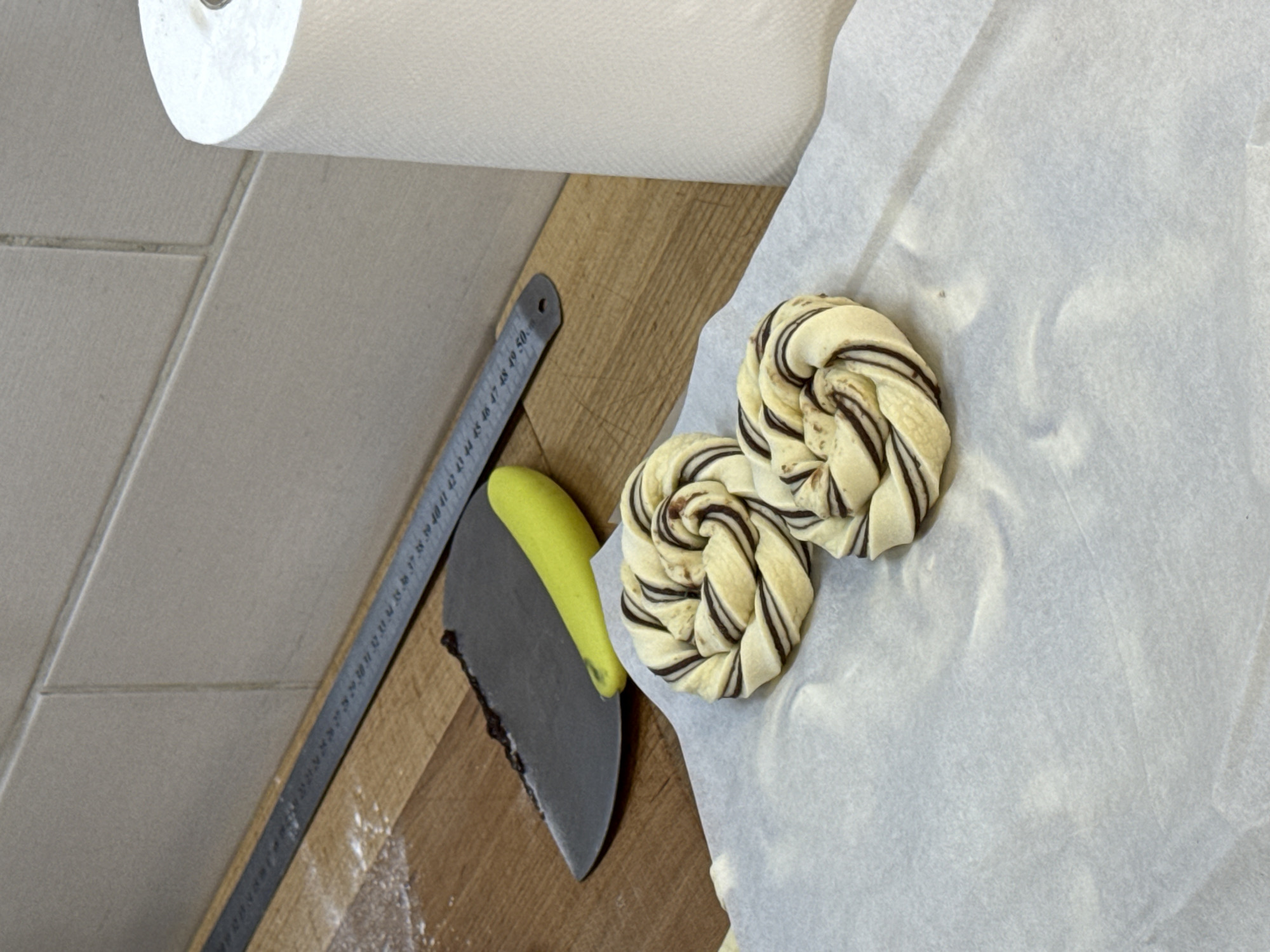 The image shows a kitchen setting with a roll of paper towels, a metal ruler, a dough scraper with a yellow handle, and two unbaked pastries on a sheet of parchment paper, all resting on a wooden countertop. The countertop is adjacent to a section of tiled wall. There are no people visible in the image. The pastries are swirled with a dark filling, likely chocolate or a similar ingredient. The location in the city of Vracar Urban Municipality, Serbia cannot be confirmed using the objects and setting as depicted in the image.
