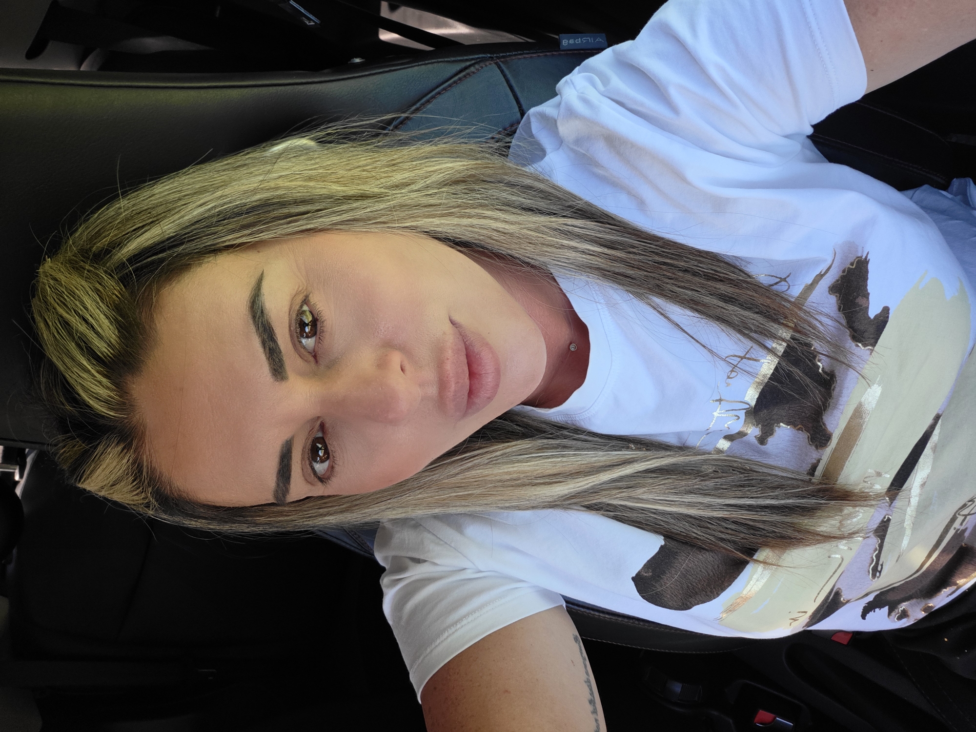 An outdoor, eye-level, medium shot captures a fair-skinned woman with long, blonde and brown highlighted hair, styled down her shoulders. She is looking at the camera with a neutral expression. She is wearing a white t-shirt with a abstract, gold, black, and white design on it, a silver necklace, and a single silver piercing below her lip. The woman is sitting in the driver's seat of a car, suggested by the black leather seat and the seatbelt buckle visible on the left side of the frame. The interior of the car is dark, with no other occupants visible. The lighting suggests it is daytime. The background is dark and indistinct. There are no landmarks or identifying features to indicate a specific location within Carpi, Italy, or anywhere else.