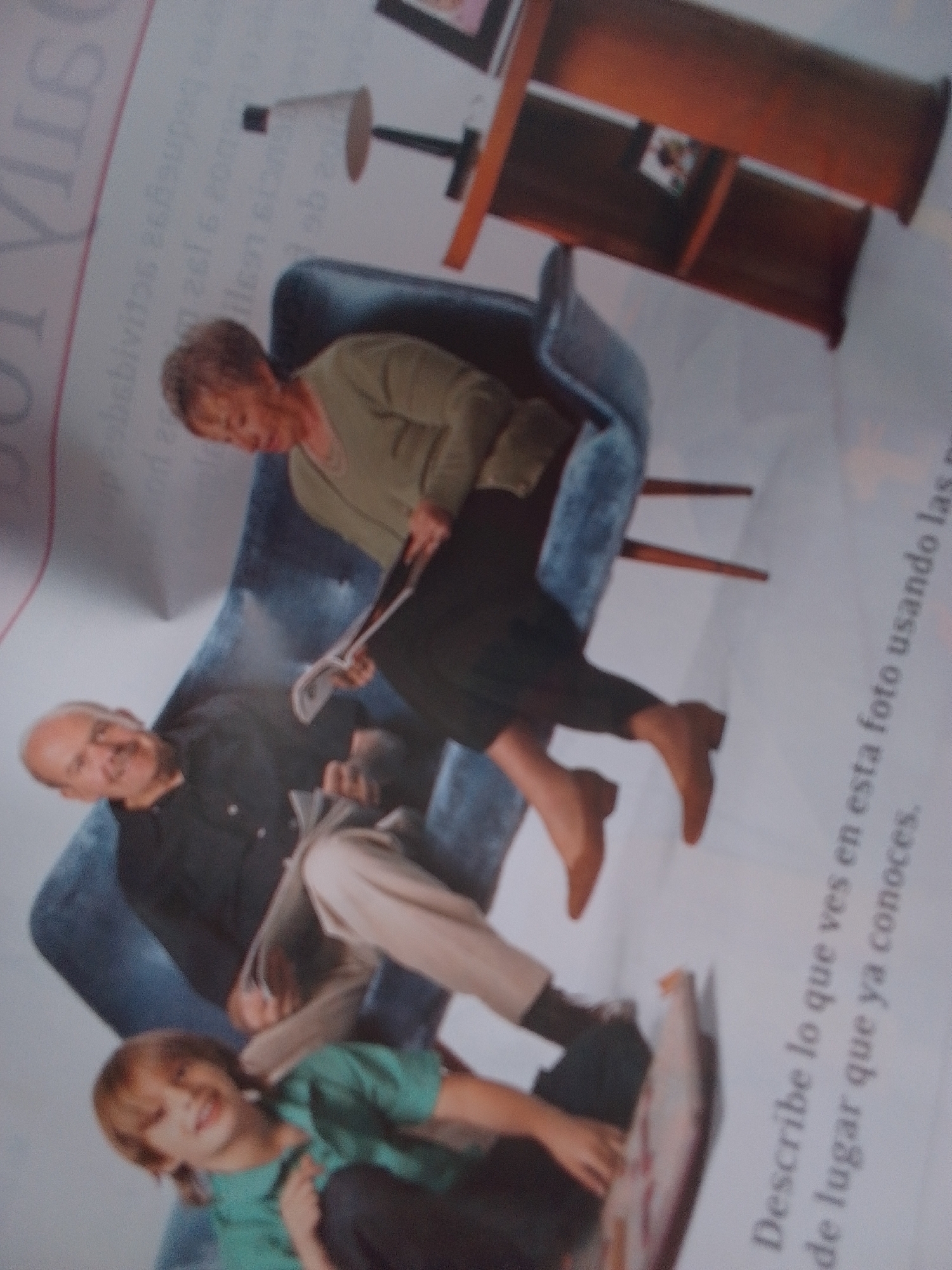 The image depicts a family scene inside a home. An elderly woman with short brown hair, wearing a green sweater and dark pants, is seated on a blue sofa, holding a newspaper. Next to her, an elderly man with short gray hair and a black shirt is also seated on the sofa, with his head tilted back. In front of the sofa, a young boy with brown hair, wearing a green t-shirt, is lying down, looking towards the camera with a smile. The background shows parts of a wooden shelf and a lamp. The image is taken from a slightly elevated angle, with the subjects oriented towards the lower left of the frame. There is no indication or information about the location being the city of La Tagua, Venezuela.