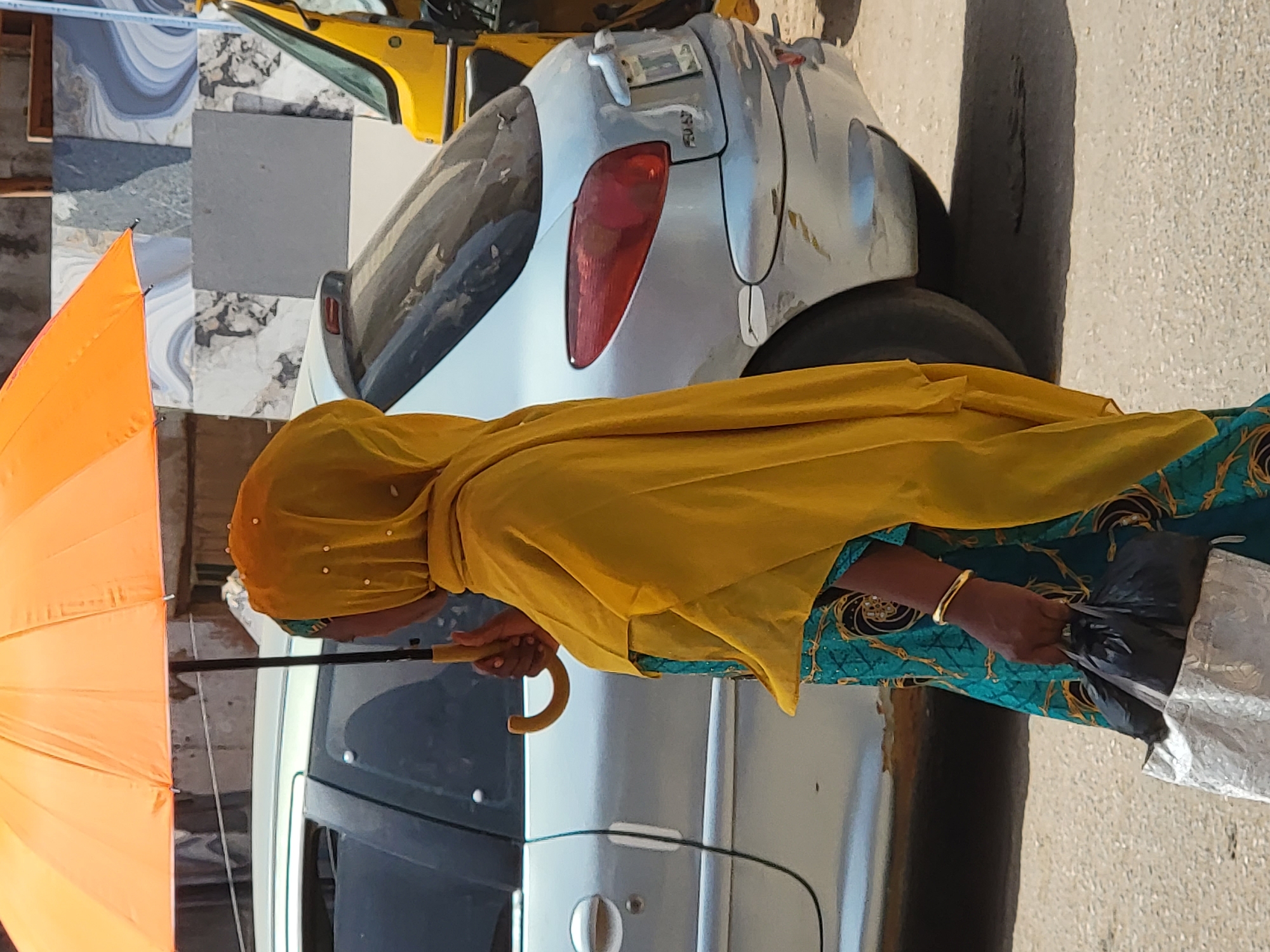 A woman is standing on a street in Maiburiji, Nigeria. She is wearing a yellow headscarf, a blue and green patterned dress, and a gold bracelet. She is holding an orange umbrella in her left hand and a black plastic bag in her right hand. Behind her is a silver car parked on the side of the road. In the background, there is another vehicle (yellow) partially visible, and a building with patterned tiles. The street appears to be paved.