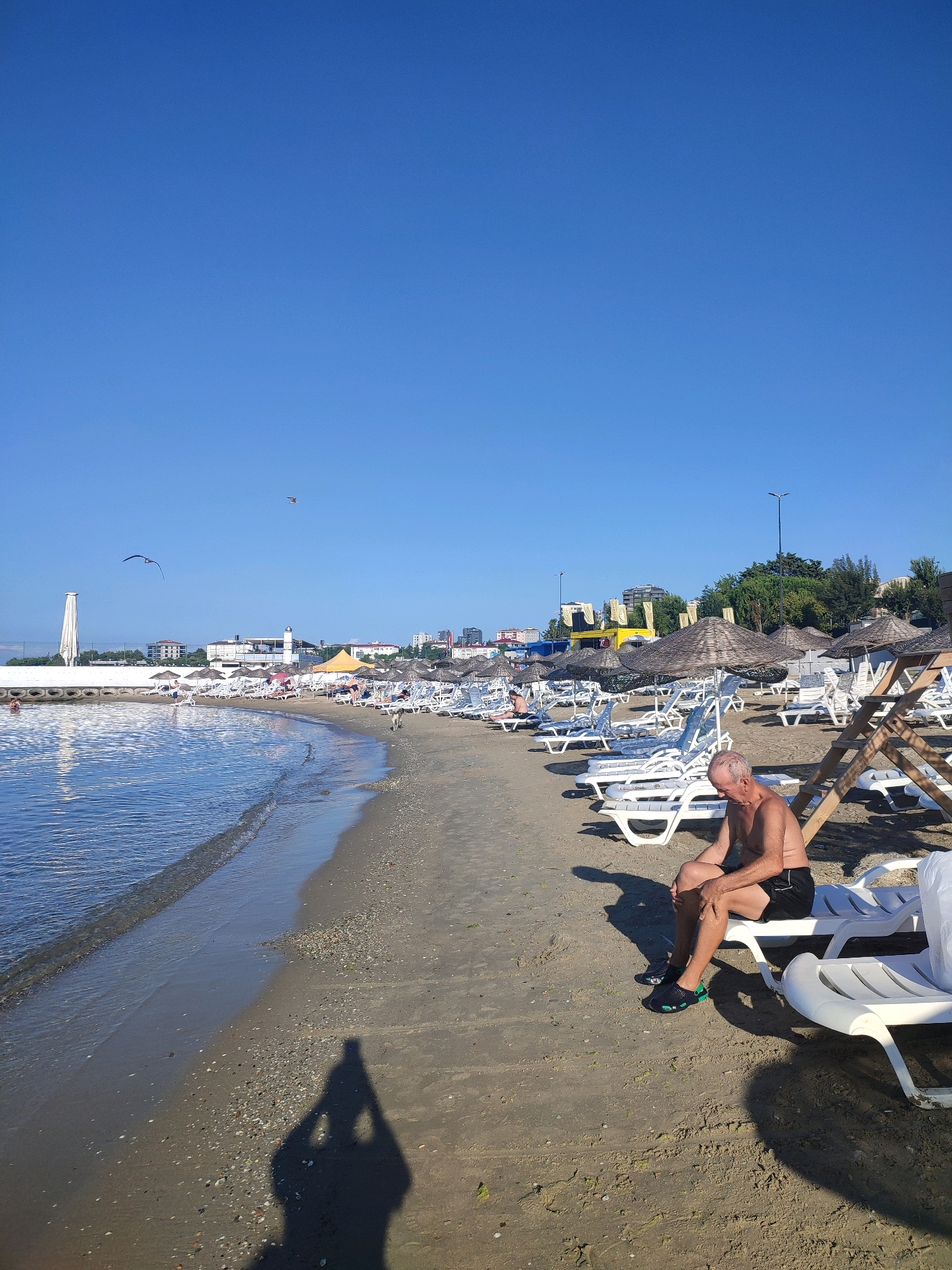 A man is sitting on a white plastic lounge chair on a sandy beach. He is shirtless and wearing black shorts and black and green shoes. He is looking down at his legs. The beach has rows of empty lounge chairs and thatched umbrellas. The sea is to the left of the frame, with gentle waves rolling onto the shore. In the background, there are buildings, trees, and a tall white structure. The sky is clear and blue. This scene appears to be a public beach in Kadıköy, Turkey, likely during the daytime.
