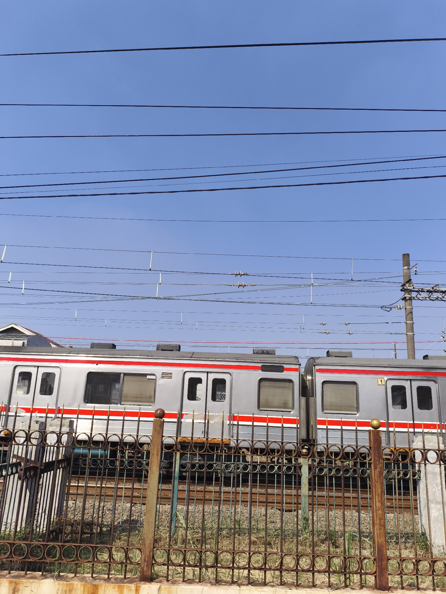 The image depicts a static, brightly lit outdoor scene on a clear day in Depok, Indonesia, featuring a silver-colored electric commuter train partially obscured by a rusted metal fence. Approximately the upper two-fifths of the frame consists of a cloudless, bright blue sky traversed by multiple horizontal black overhead power and communication lines. Several white or light-colored vertical insulators/hangers are suspended from these wires. On the far right, a brown utility pole with metal bracing extends upwards, supporting additional overhead infrastructure.
In the mid-ground, a commuter train, likely an electric multiple unit (EMU) consistent with KRL Commuterline operations, is positioned on railway tracks. The train cars are predominantly silver with a prominent horizontal red stripe running below the windows. Rectangular windows and doors are visible on the train's side; one window on the left-center car shows a partially drawn blind. A yellow rectangular sticker bearing the number "1" is affixed to a door on the rightmost visible car. The roof of the train shows several black components, possibly air conditioning units. The train appears stationary.
The foreground is dominated by a dark, heavily rusted metal fence with repeating circular patterns along its top and bottom horizontal bars. Several vertical support posts, varying in color from rusted brown to faded green, anchor the fence. One prominent brown post on the right features a spherical finial. The fence rests upon a concrete base, partially visible at the very bottom of the frame, exhibiting rust stains. Behind the fence, the railway tracks are visible, comprising parallel rails resting on sleepers (ties) within a bed of gravel ballast. Sparse patches of dry grass and weeds grow between the fence and the tracks. No individuals are present in the scene.