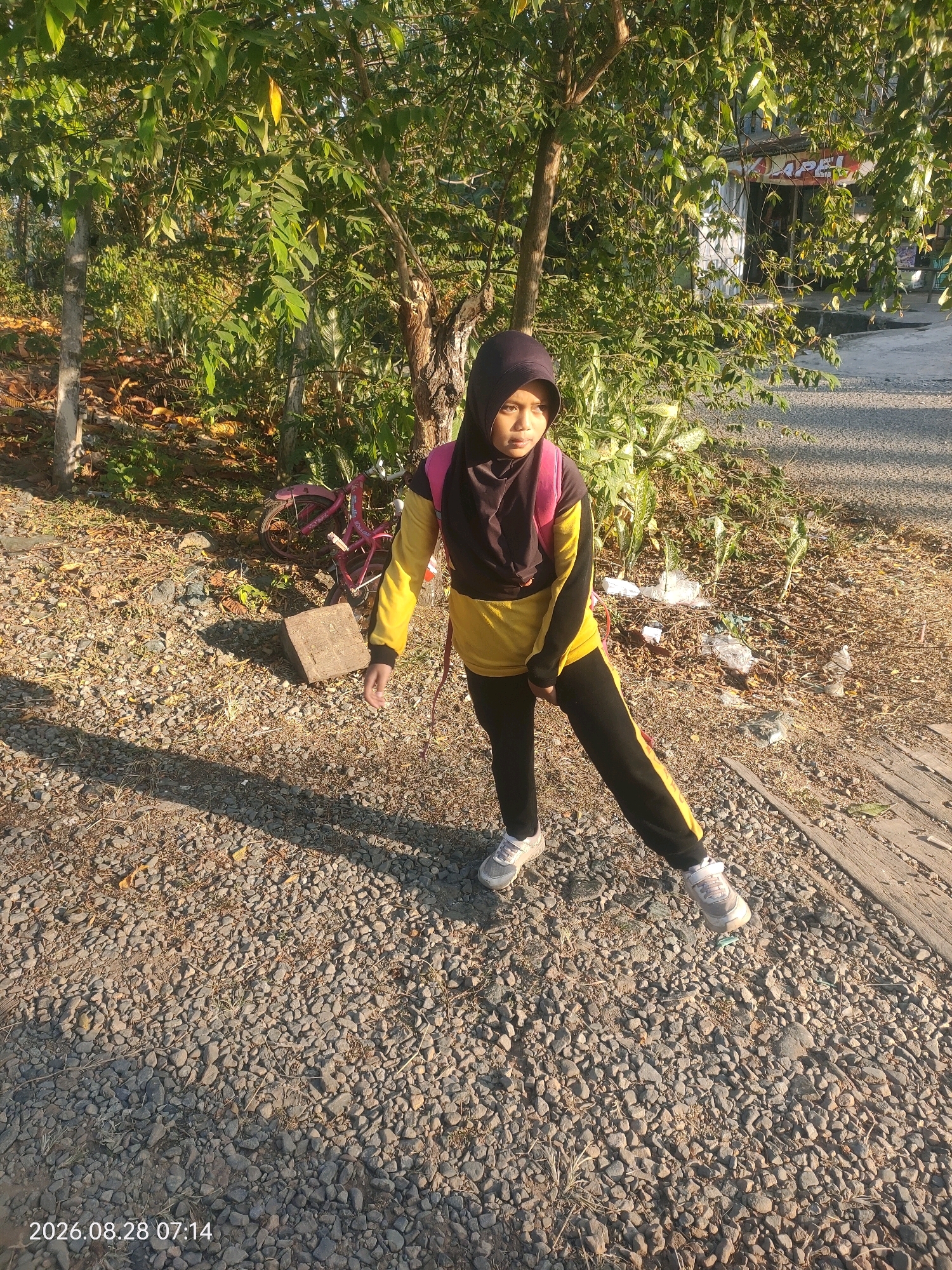 A young female wearing a black hijab and a yellow and black athletic outfit, stands outdoors on a gravel path next to a dirt embankment with sparse vegetation. She has a pink backpack on and is looking to her left, with her right leg extended forward. Behind her, partially obscured by trees, is a pink bicycle and a concrete block. In the background, a building with signage is visible, and the ground is covered in small stones and dry foliage. The scene is illuminated by sunlight, casting long shadows across the path. The timestamp on the image reads 2026.08.28 07:14.