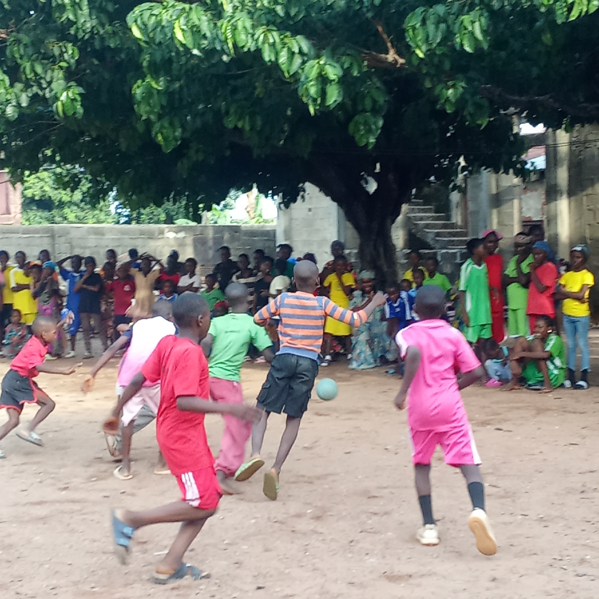 Akwanga, Nigeria: A group of children play soccer in a sandy, open area under the shade of a large tree. Spectators, mainly children and some adults, stand along the periphery, watching the game. The players are dressed in various colored shirts and shorts, with one child wearing a striped shirt kicking the soccer ball. In the background is a building with exposed brick and what looks like outdoor stairs. The weather appears to be sunny.