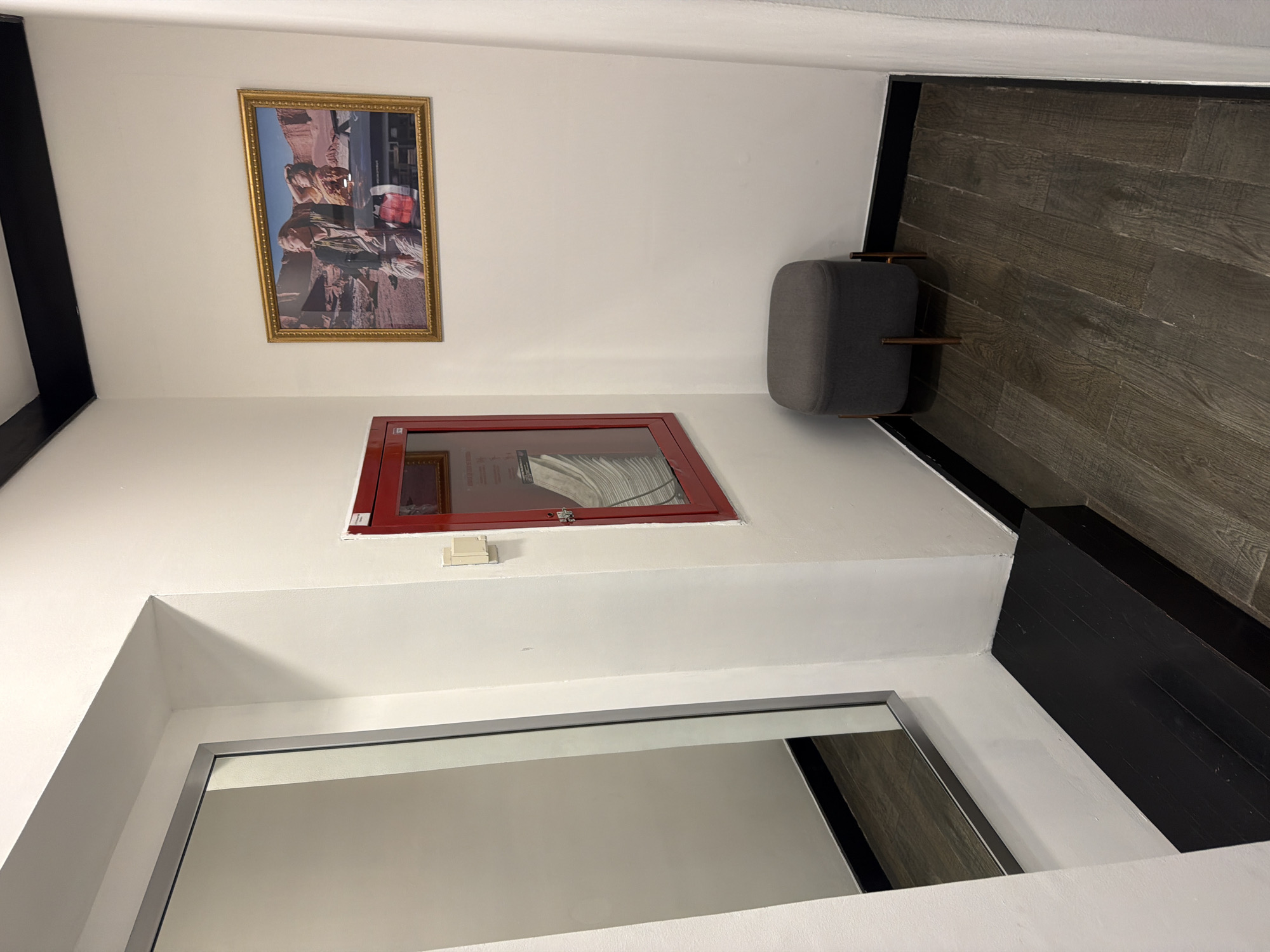The image shows a white-walled room with a dark wood-look floor and black trim. A framed picture of people in a desert landscape hangs on the wall. Below the picture is a red-framed object, likely a fire safety box. A gray ottoman sits near the corner of the room. A silver-framed mirror is positioned near the bottom of the image, reflecting part of the room. The location is possibly in the city of Puebla, Mexico, but there are no visible elements to confirm this.