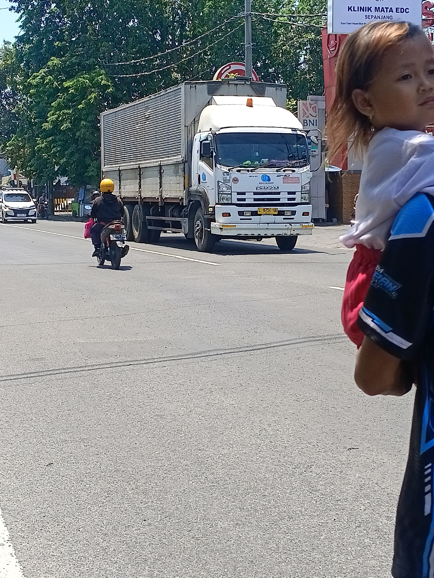 The image shows a street scene in Kalijaten, Indonesia. In the foreground, a young child is being carried on someone's shoulders. The child's face is visible, looking to the right. The person carrying the child is wearing a dark shirt with blue and white accents.
In the background, a large white truck with a silver cargo container is driving on the road. The truck has "ISUZU" branding visible on the front. A person wearing a yellow helmet and a dark jacket is riding a motorcycle in front of the truck, also traveling on the road. To the left of the truck, a white car is also visible on the road.
The environment appears to be a sunny day with green trees lining the background. There are also buildings visible, including one with signage that says "KLINIK MATA EDC" and "SEPANJANG," and another with "BNI" branding. The road is asphalt, and there are road markings visible.