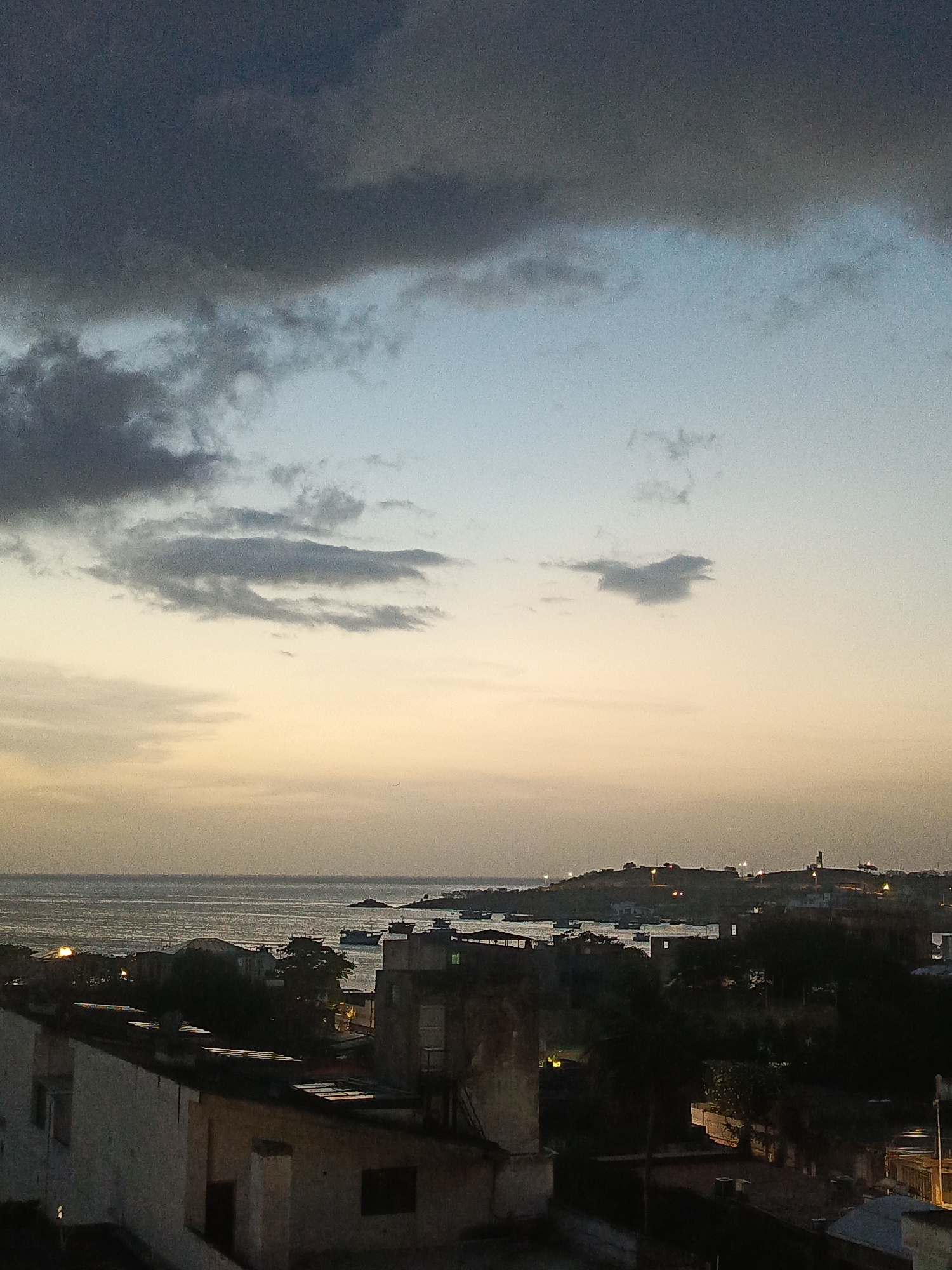 The image depicts a twilight view of Juan Griego, Venezuela. In the foreground, several buildings with stark white and weathered facades are visible, some with visible window openings and architectural details. The rooftops are predominantly dark, suggesting a low-light condition. Beyond the buildings, a body of water, likely the sea, stretches towards the horizon. Several small boats are scattered on the water, some appearing to be anchored. In the distance, a hilly coastline is lined with scattered lights, indicating a settlement. A prominent structure resembling a lighthouse or tower stands on one of the higher points of the coastline. The sky is a gradient of colors, transitioning from a pale yellow near the horizon to a soft blue and then to dark, heavy clouds in the upper portion of the frame. The overall atmosphere is serene and suggestive of the approaching night.