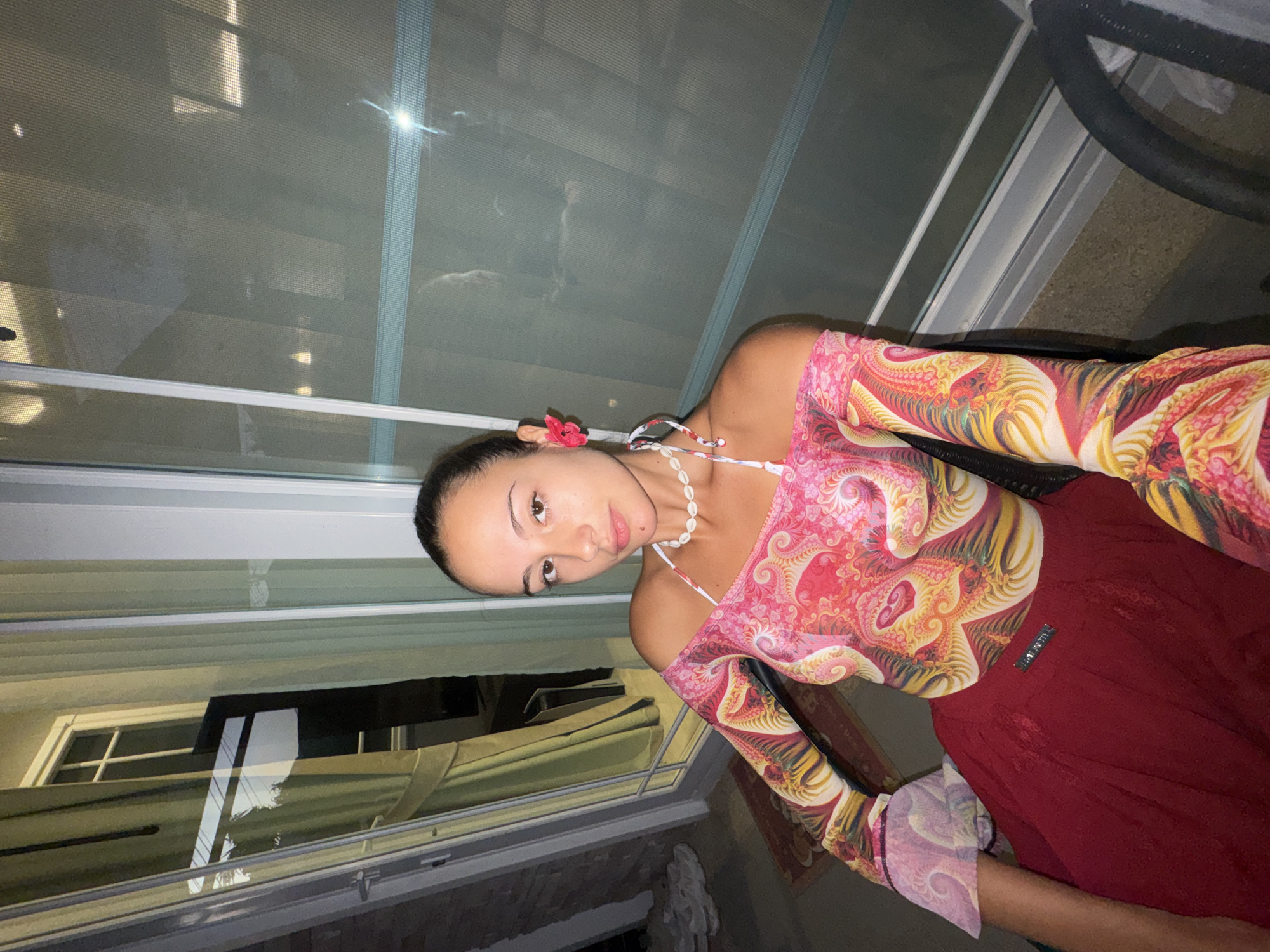 A woman poses indoors near a window, facing the camera at an angle. She has brown hair pulled back, fair skin, and wears a red flower in her hair, a shell necklace, and a pink and yellow long-sleeved top. She wears a maroon skirt. The window behind her shows a view of what appears to be another building, potentially in Chalong, Thailand, given the request. The lighting is dim.