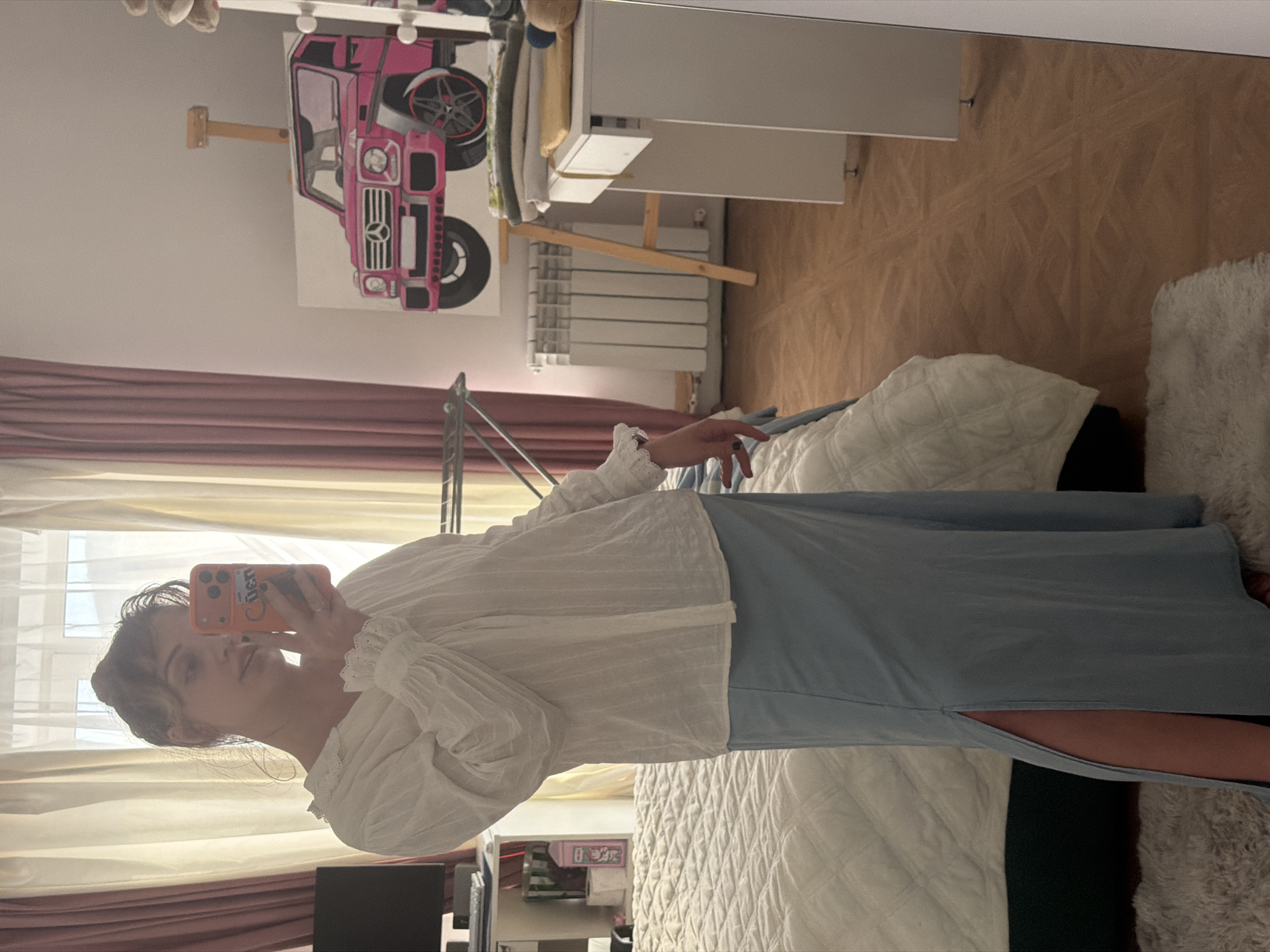 A young woman is depicted taking a mirror selfie in a well-lit room, likely a bedroom or dressing area. She is standing and looking into her phone, held up to capture her reflection. She wears a white, intricately textured blouse with ruffled details on the collar and voluminous, gathered sleeves. Her lower body is clad in a light blue long skirt featuring a high slit on the right side, revealing her leg.
The room behind the woman (and reflected in the mirror) features a large painting of a pink Mercedes-Benz G-Wagen (G-Class) on a light-colored wall. Below the painting, a white radiator and a white dresser or cabinet are visible. The floor is light brown, and on the right side of the frame, a bed with white quilted bedding and a fluffy white throw is partially visible. Pink and sheer white curtains hang over a window on the left, letting in natural daylight. A metal clothes rack is also partially visible on the left.
The woman's smartphone is encased in a vibrant orange cover, which displays partially visible text that appears to read "Cüden". The overall ambiance of the scene is casual and bright, typical of a daytime indoor setting. The location context is Большая Шильна, Russia.