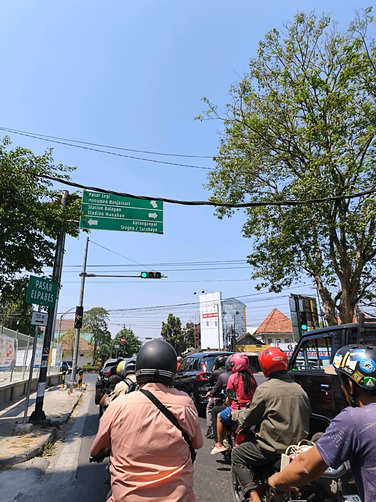 The image shows a traffic scene on a sunny day in the city of Surakarta, Indonesia, likely near the intersection of Jalan Slamet Riyadi and Jalan Gajah Mada. In the foreground, several people are riding on motorcycles, viewed from behind. One individual in a peach-colored shirt has a black helmet and a black strap across their shoulder. Another rider wears a red helmet and a green jacket, while a third is in a pink helmet and a pink shirt. Further back, a person in a black helmet with stickers is visible. The vehicles are stopped at a traffic light, which is illuminated with a green light.
Above the intersection, green directional signs are mounted on poles and wires. One sign indicates "Pasar Legi," "Monumen Banjarsari," "Stasiun Balapan," and "Stadion Manahan" with arrows pointing to the left and right. Another sign reads "Karanganyar," "Sragen / Surabaya" with an arrow pointing right. A smaller green sign on a pole to the left reads "PASAR ELPABES" with an arrow pointing left and a distance marker "150 m."
The environment includes a clear blue sky, abundant foliage from a large tree on the right side of the image, and several buildings in the background. Power lines are strung across the sky, typical of an urban setting. The road is asphalt, and there is a sidewalk with some greenery. The overall impression is of a busy urban intersection with moderate traffic flow.