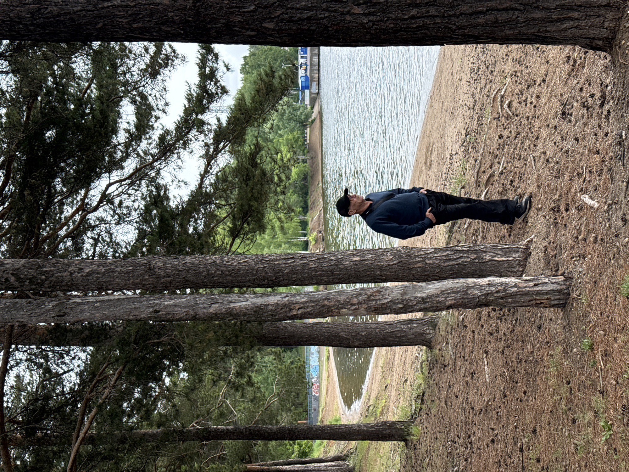 The image captures an outdoor scene in the city of Chaikovsky, Russia, depicting an adult male on a tree-lined sandy shore next to a body of water. The man, positioned right of center, is observed in a three-quarter profile, looking to his left. He is wearing a dark blue long-sleeved garment, black pants, a black baseball cap, and eyeglasses. A dark strap, possibly from a bag, crosses his left shoulder. His hands are placed in his pockets.
The ground in the foreground is covered extensively with reddish-brown pine needles and numerous small pine cones. Several tall, straight pine tree trunks, with dark, textured bark, are visible, ascending vertically and framing the scene. Their branches, bearing green needles, form a partial canopy overhead, with glimpses of a pale, overcast sky.
A sandy beach, lighter in color than the pine-needle-covered ground, forms a narrow strip along the water's edge. The body of water, likely a lake or river, extends from the midground, curving towards the left side of the frame. Its surface is calm with light ripples, reflecting the sky and surrounding vegetation.
Across the water, in the left background, a concrete or stone embankment with a blue railing is visible, featuring multi-colored graffiti on its lower section. Further along the opposite shore to the right, several small, boxy blue structures are discernible, potentially small buildings or kiosks, situated near what appears to be a pier or dock. Beyond the far shore, a dense expanse of green forest dominates the background, with a metallic utility tower faintly visible above the distant treeline. The sky is uniformly light gray, indicating an overcast condition.