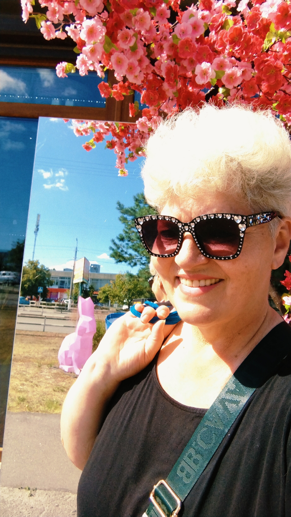 A woman with blonde, permed hair and oversized sunglasses with rhinestones poses for a selfie outdoors. She is wearing a black tank top and a green strap with text is visible across her chest. Behind her, to the left, is a reflective surface, possibly a large mirror or window, which shows a reflection of the sky, trees, buildings, and a pink sculpture resembling a rabbit. In front of the reflective surface, a pink sculpture of a rabbit is visible. Red and pink artificial flowers are arranged above the woman's head. The background suggests an urban environment with buildings, trees, a street, and a traffic sign. The sky is clear and blue with some clouds. The location is identified as Horishni Plavni, Ukraine.