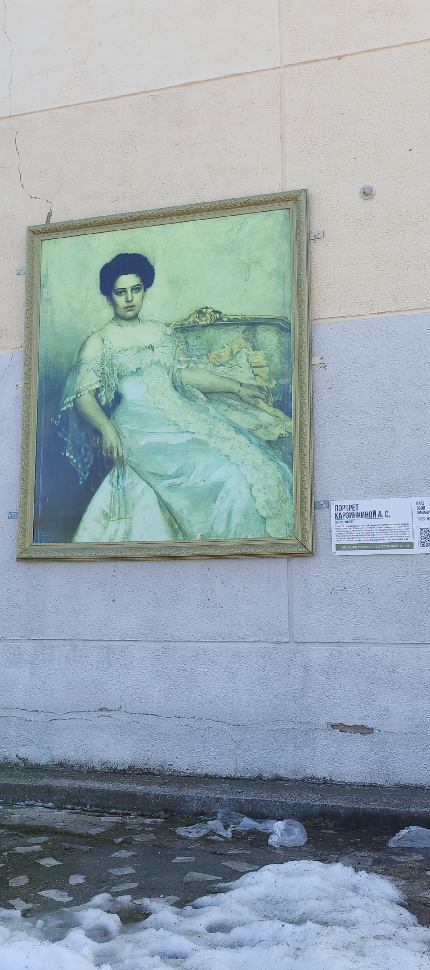 A framed portrait of a woman hangs on a wall outside. The portrait is likely a print or reproduction as it is mounted outdoors. The portrait's informational plaque indicates this is a portrait of A.S. Karzinkin and is located in Chaikovsky, Russia. Snow and ice are visible at the base of the wall in the foreground. The wall appears to be part of a building. The conditions seem cold and possibly late winter or early spring.