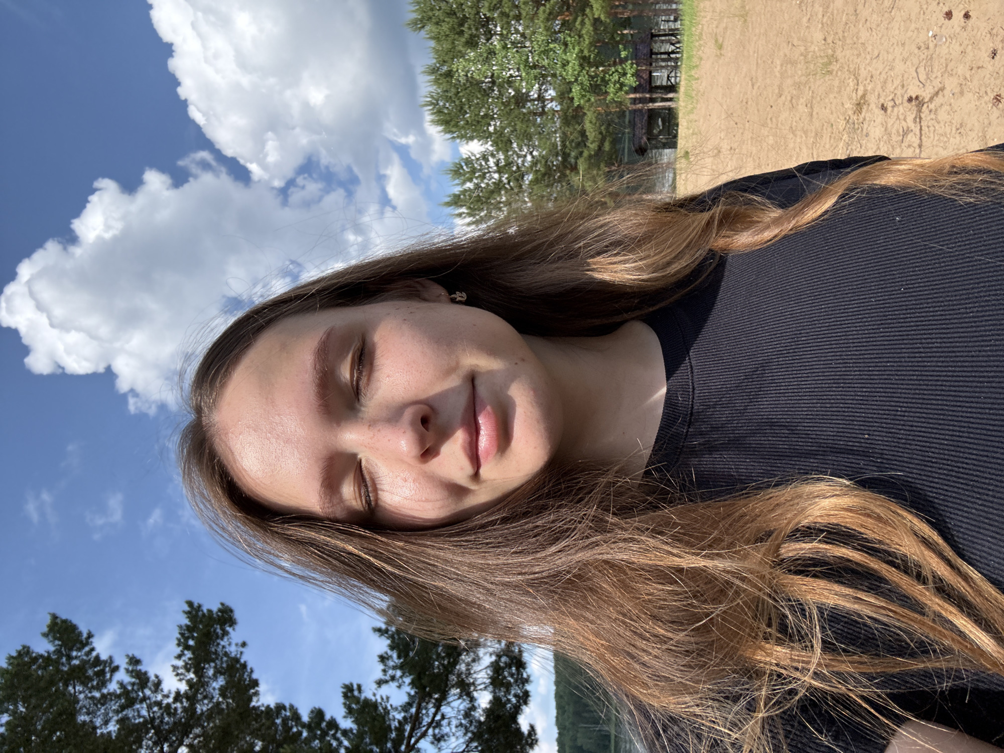 A young Caucasian woman with long, wavy brown hair and closed eyes is taking a selfie outdoors. She is wearing a black, ribbed t-shirt and a small, silver stud earring in her left ear. Her face is turned slightly towards the camera, with a faint smile. The background consists of a sandy beach, a body of water, a line of pine trees, and a wooden gazebo. The sky is blue with fluffy white clouds. The lighting suggests it is daytime, likely mid-morning or afternoon, with sunlight illuminating the scene. The location appears to be a park or recreational area, possibly near a lake or river within the city of Chaykovsky, Russia.