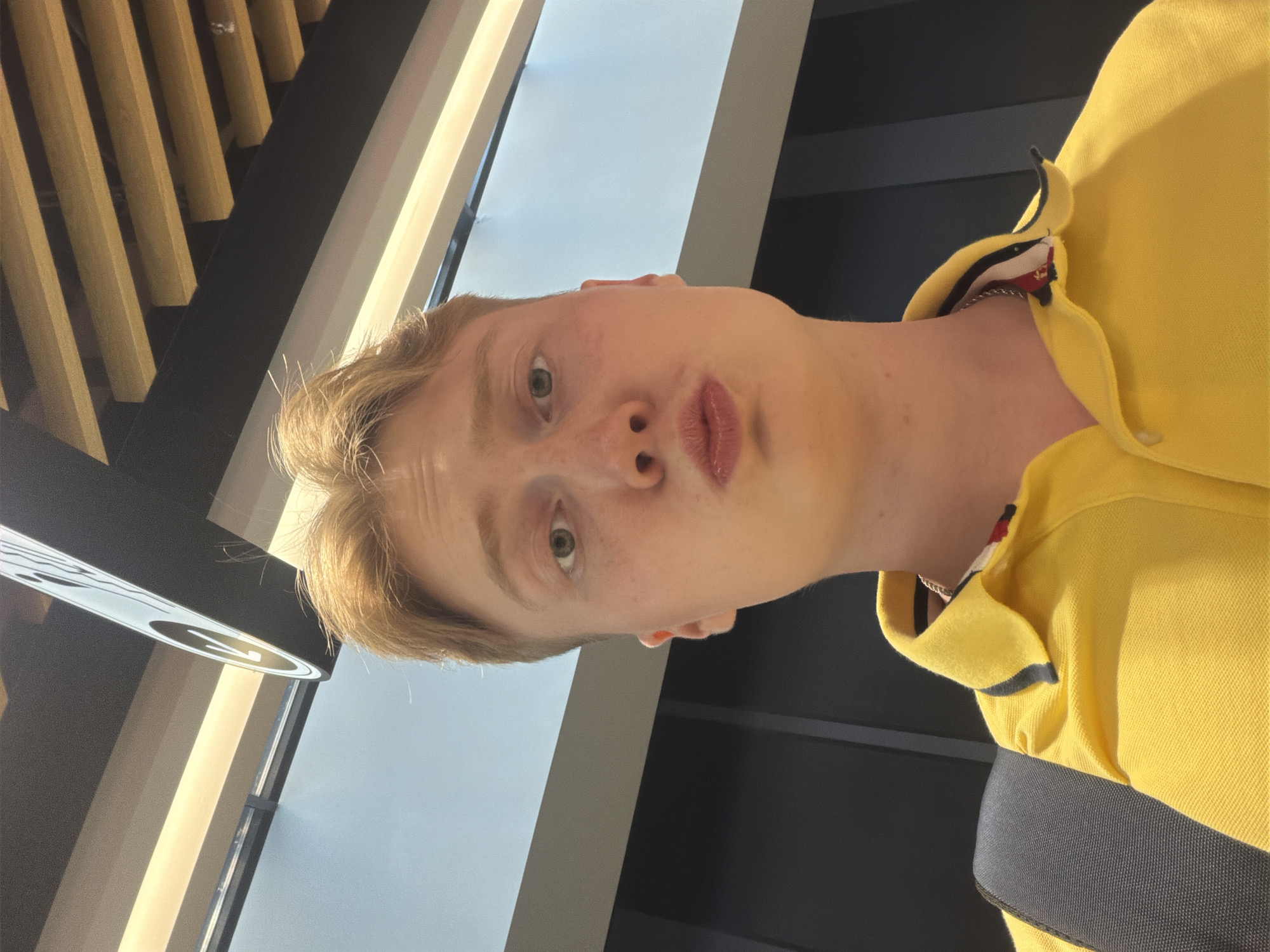 A selfie of a young man with fair skin, light brown hair, and green eyes is taken in an indoor environment. He is wearing a yellow polo shirt with a red, white, and black trim on the collar. His lips are pursed. Behind him, a black backpack is visible. The background features modern architectural elements with a dark ceiling, light-colored panels, and yellow wooden beams. There is also a black wall with vertical gray lines. A sign with a white arrow on a black circle is also present in the upper part of the image. The lighting appears to be artificial, coming from overhead fixtures. The overall scene suggests an indoor public space, possibly a transportation hub or a modern building in a city like Kyiv, Ukraine, given the context.
