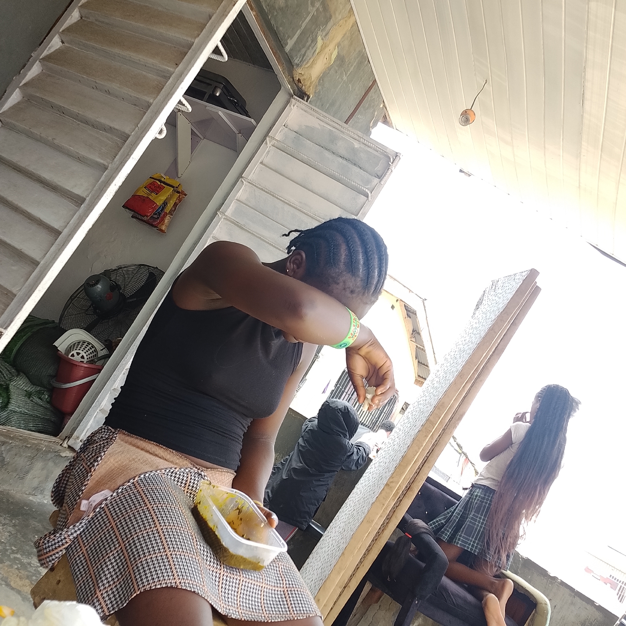 In Ado, Nigeria, an outdoor scene unfolds with a focus on a young woman seated on a stool, her face partially obscured as she wipes her eyes with her arm. She wears a black tank top and a plaid skirt. A clear plastic container of food sits beside her. In the background, another young woman with long braided hair, wearing a white t-shirt and a plaid skirt, is visible near a dark seating structure. The environment includes a partial view of stairs, a fan, bags of items, and corrugated metal roofing, suggesting a utilitarian, possibly residential or commercial outdoor space. The lighting indicates it is daytime.