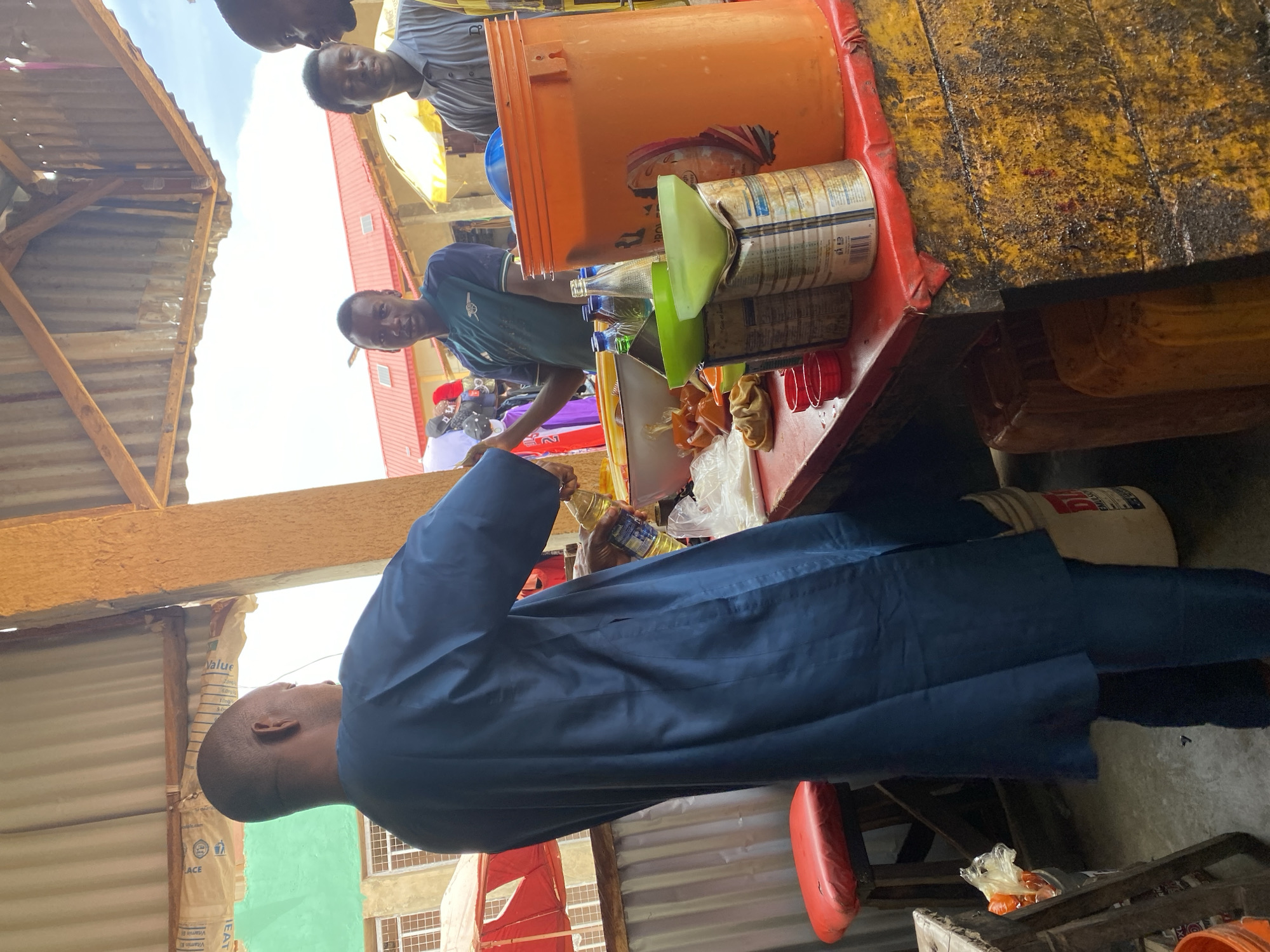 In Kafanchan B, Nigeria, the image captures an outdoor marketplace scene. A man in a dark blue garment stands near a food stall. He is holding a bottle of cooking oil. On the stall, there are various items including an orange bucket, tinned goods, bottles, a green funnel, small red cups, and bagged items that appear to contain food. Two other men are positioned behind the stall. The surrounding environment consists of corrugated metal structures, suggesting a makeshift or informal marketplace setting.