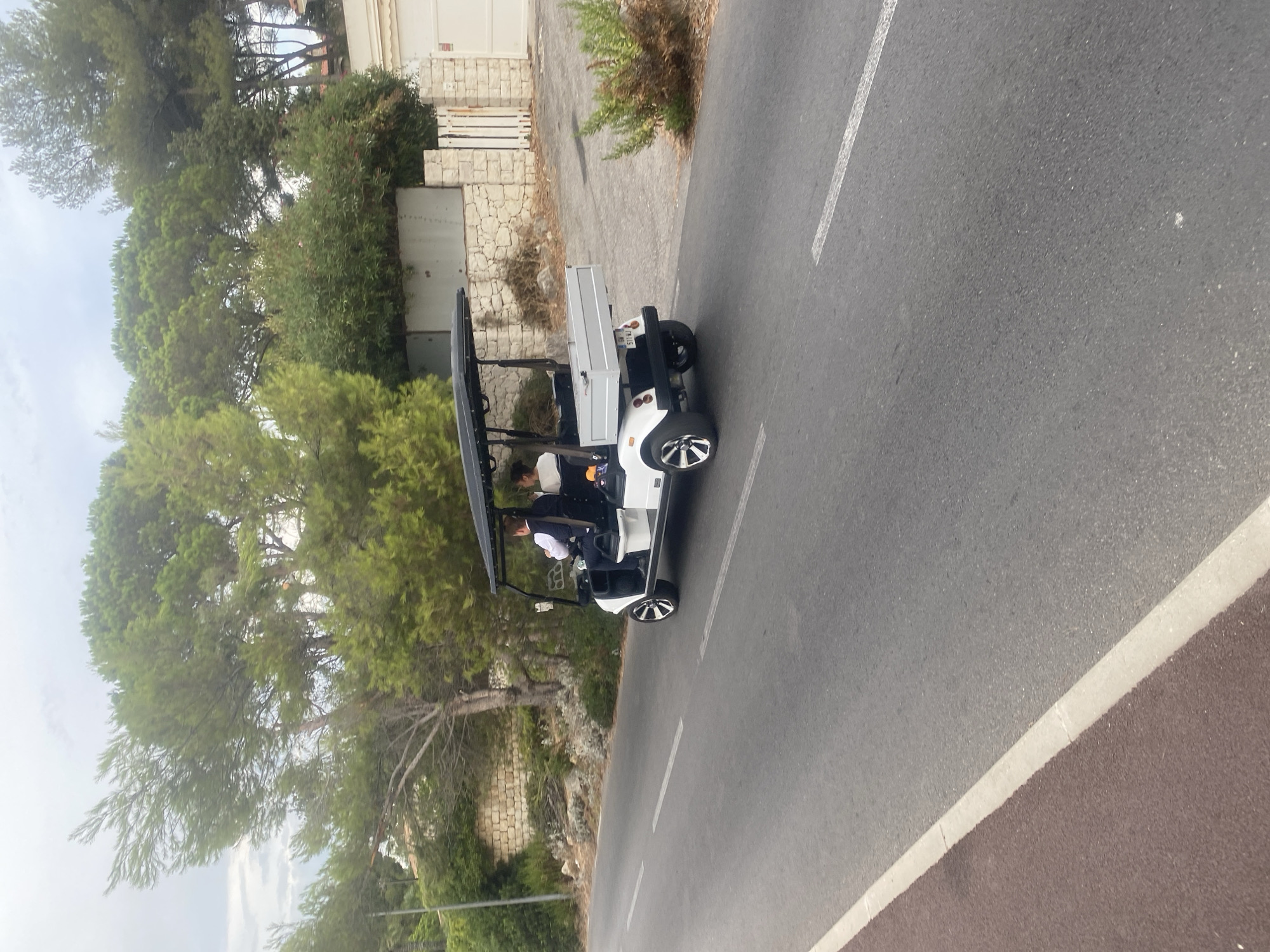 In the city of Antibes, France, a white golf cart with a black canopy is driving on an asphalt road with white lane markings. Two individuals are seated in the golf cart; the driver, wearing a dark vest and white shirt, is operating the vehicle, while a passenger, wearing a white shirt, is seated to the driver's right. The golf cart has a small silver cargo bed attached to the rear. The road is flanked by a red-colored paved shoulder on the left and a stone wall with foliage and trees on the right. In the background, there are more trees and buildings, including a white fence and a garage door. The sky is overcast.