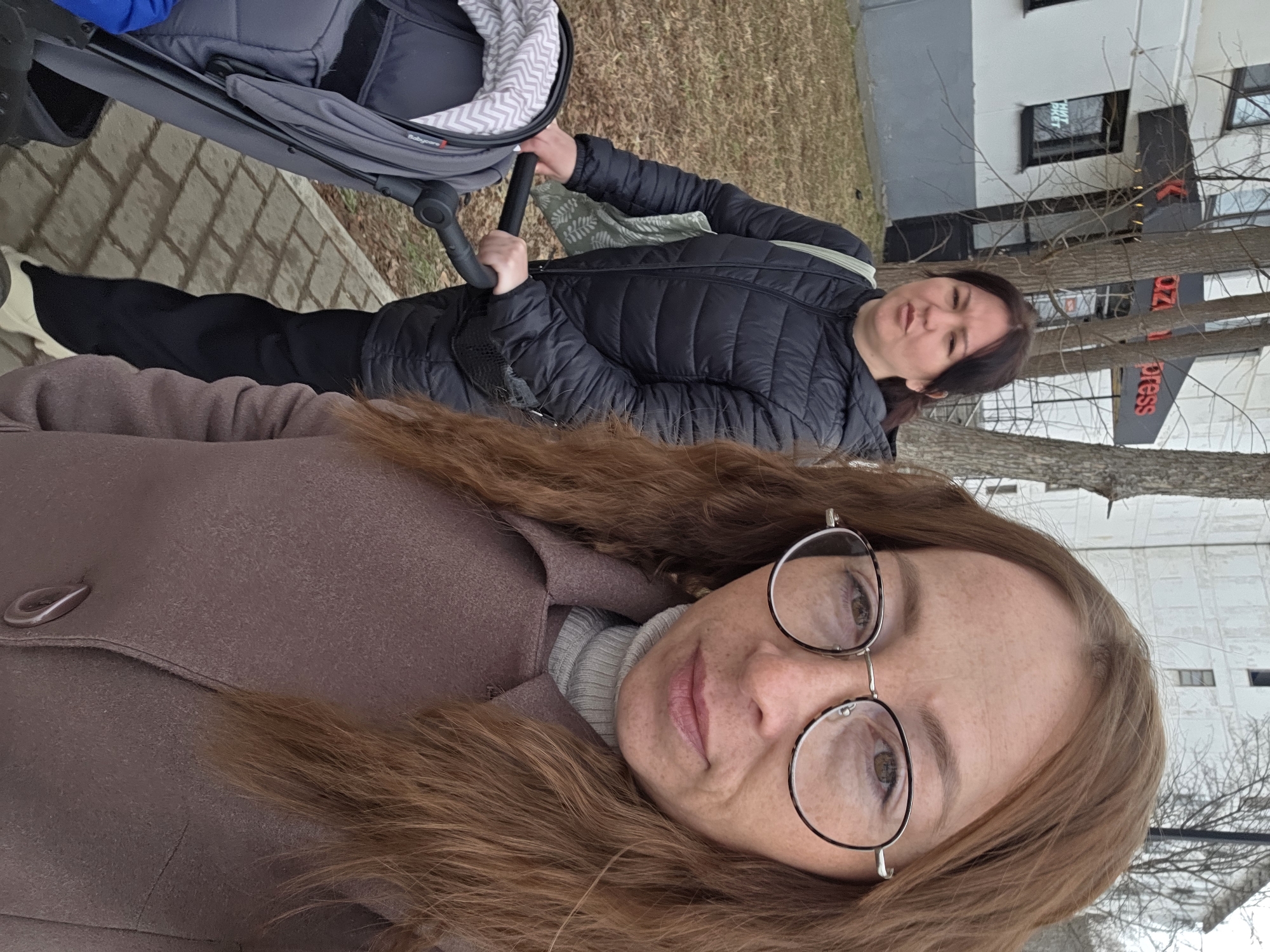 The scene depicts two women on a paved sidewalk next to a commercial building in Chaikovsky, Russia, on an overcast day.
In the immediate foreground, a woman with long, wavy reddish-brown hair, visible freckles, and round-rimmed glasses is taking a selfie, looking directly at the camera. She is wearing a light grey ribbed turtleneck and a collared brown/mauve-colored coat.
Behind her and slightly to the left, another woman with dark brown hair, pulled back, is pushing a grey baby stroller. The stroller has a black handle and a lighter grey and white chevron pattern on its interior fabric; the brand "Babycare" is partially visible. A blue object is seen inside the stroller. The woman is wearing a black quilted puffer jacket, black pants, light-colored sneakers, and is carrying a light green tote bag with a leaf pattern on her left shoulder, along with a small black pouch strapped across her hip. Her gaze is directed towards the left of the frame, not at the camera.
The background features a multi-story building, predominantly white with grey sections on its ground floor. This building displays a black awning with red text "az Express" (likely part of "KazanExpress") and a small sign below it reading "НИТ МАРКЕТ" (NIT Market). Further back and to the right, another taller, light-colored building is visible. Several bare-branched deciduous trees are scattered throughout the background, indicating a cooler season. The ground to the left of the sidewalk consists of dry, sparse grass and dirt.