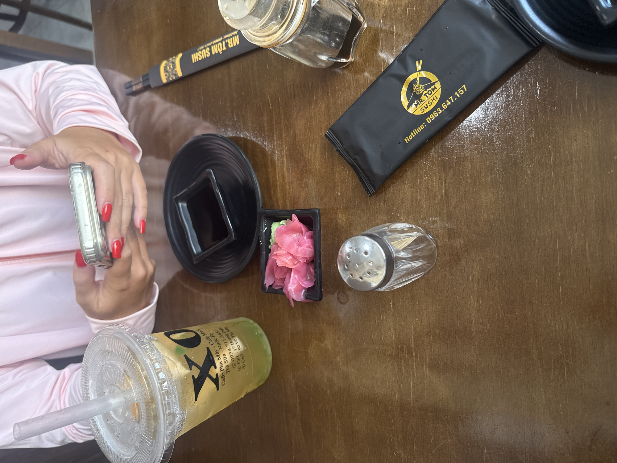 A person with red-painted fingernails is holding a mobile phone in their hands, with a pink long-sleeved shirt on. On a wooden table, there is a clear plastic cup with a yellow beverage and ice, a black square dish with soy sauce, a small black square container with pickled ginger and wasabi, a glass salt shaker, and a black packet with chopsticks and the text "MR. TOM SUSHI" and a phone number. There is also a glass bottle containing soy sauce. The overall scene appears to be a meal setting at a restaurant. The text on the plastic cup includes Vietnamese words and a phone number, suggesting the location might be Vietnam. Based on the available information, it is not possible to precisely pinpoint the location within Nha Trang, Vietnam, but the presence of Vietnamese text on the beverage cup and the restaurant's branding indicates it is likely a local establishment.