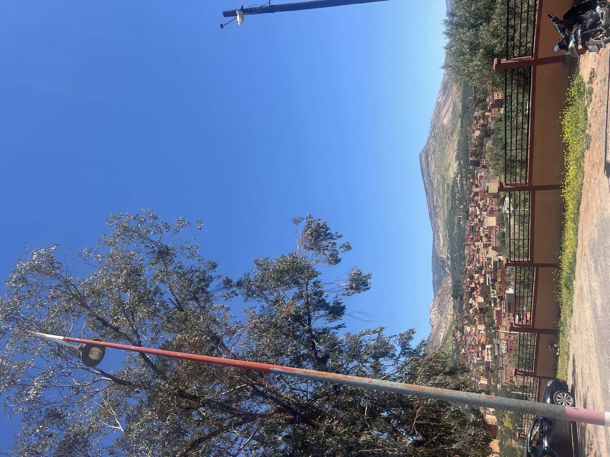 The image depicts a view in Ouaouizert, Morocco. A town nestled at the base of a mountain is visible. A red and grey pole intersects the lower portion of the frame, with a light fixture attached to its top. A tree partially obscures the view. The sky is clear and blue. A motorcycle and a car are parked next to a wall with a railing, on the right side of the frame.