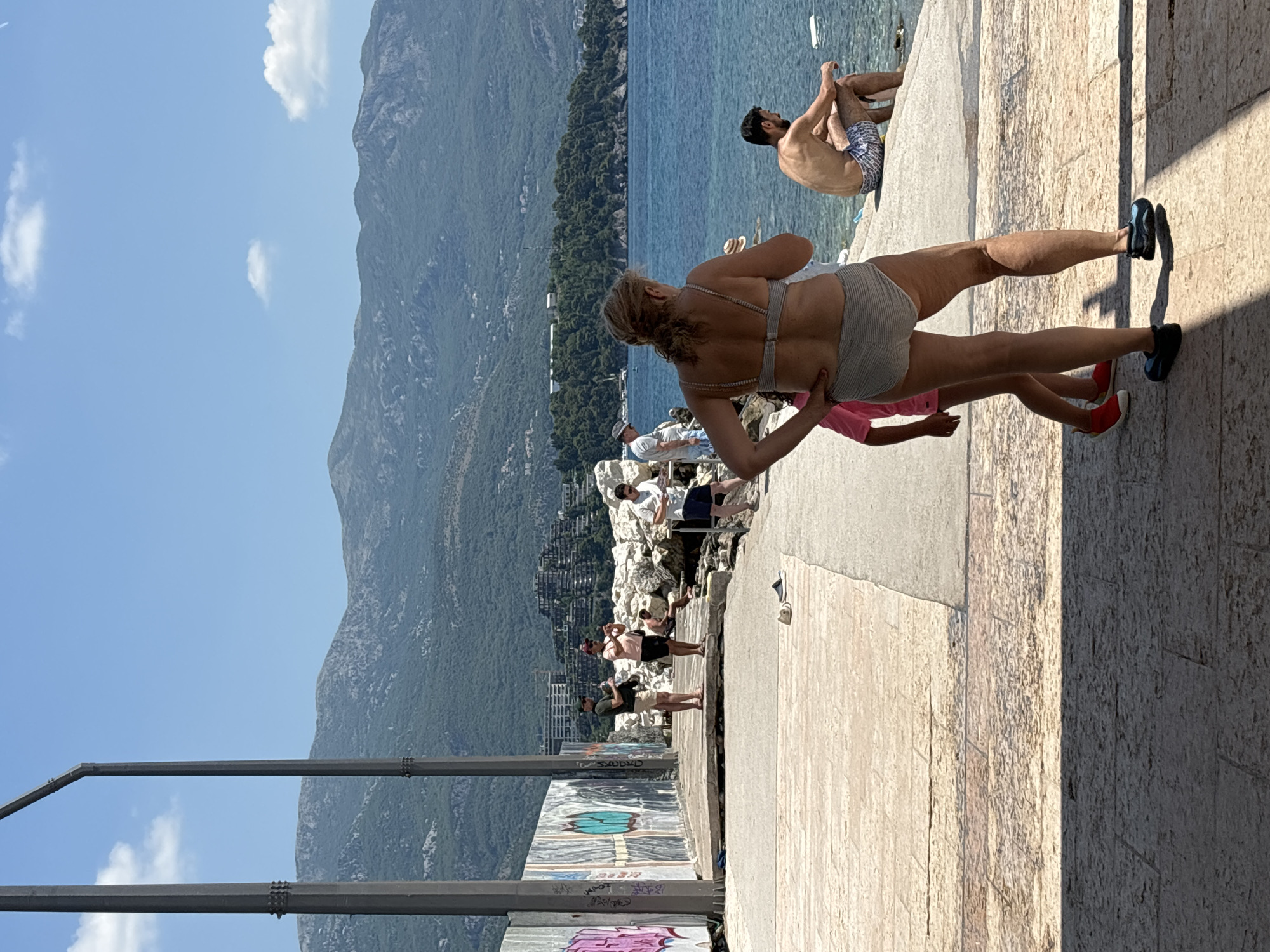 The image depicts a sunny day with clear blue skies and a few wispy clouds, overlooking a body of water and a mountainous coastline. The scene is set in Budva, Montenegro. In the foreground, a woman in a striped bikini is captured mid-stride, her muscles toned and defined. She is wearing black sneakers. To her left, another person in a pink top and red shoes is partially visible. In the middle ground, several people are gathered on a rocky area near the water. A man with dark hair is sitting on the rocks, wearing patterned swim trunks. Further back, a resort town with buildings nestled into the hillside can be seen. The water is a vibrant blue, reflecting the bright sunlight. The foreground walkway is made of light-colored stone tiles, with shadows cast by an unseen structure. There is graffiti on a concrete structure in the lower left portion of the image. The overall atmosphere suggests a leisurely summer day.