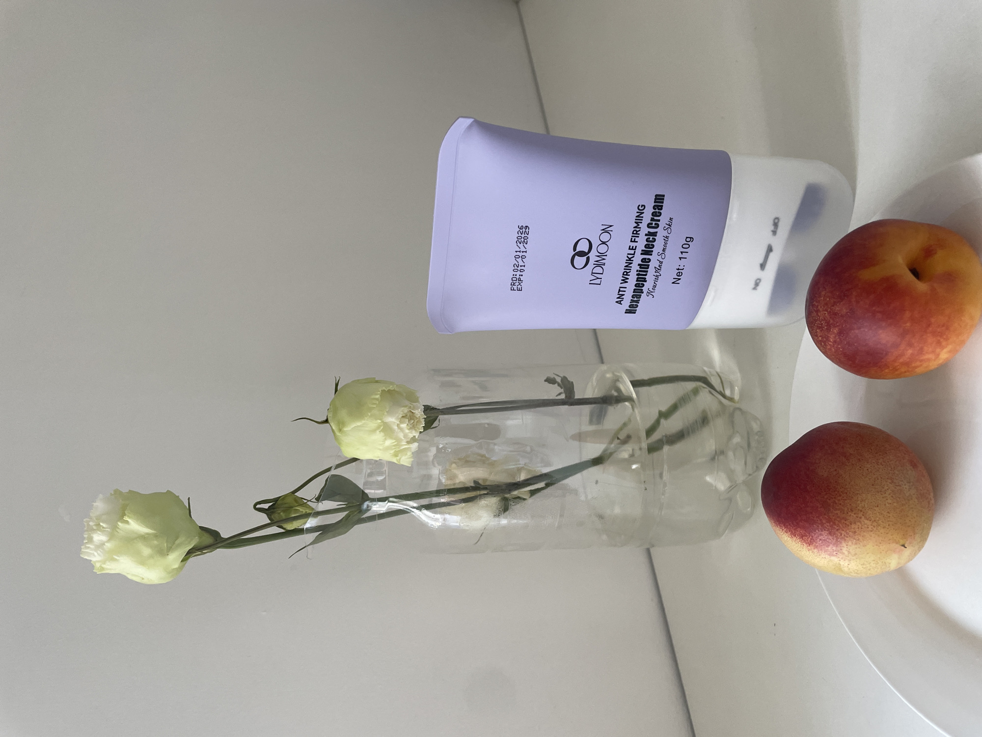A still life arrangement features a clear plastic bottle containing three wilting light green flowers with thin stems in water. To the right of the bottle is a tube of Lydymoon Anti Wrinkle Firming Hexapeptide Neck Cream, with its expiration date of 01/01/2029 visible on the label. In the foreground, two ripe nectarines rest on a white oval plate. The composition is set against a neutral, off-white background. The scene is indoors, with no specific identifiable landmarks to place it in Almaty, Kazakhstan.