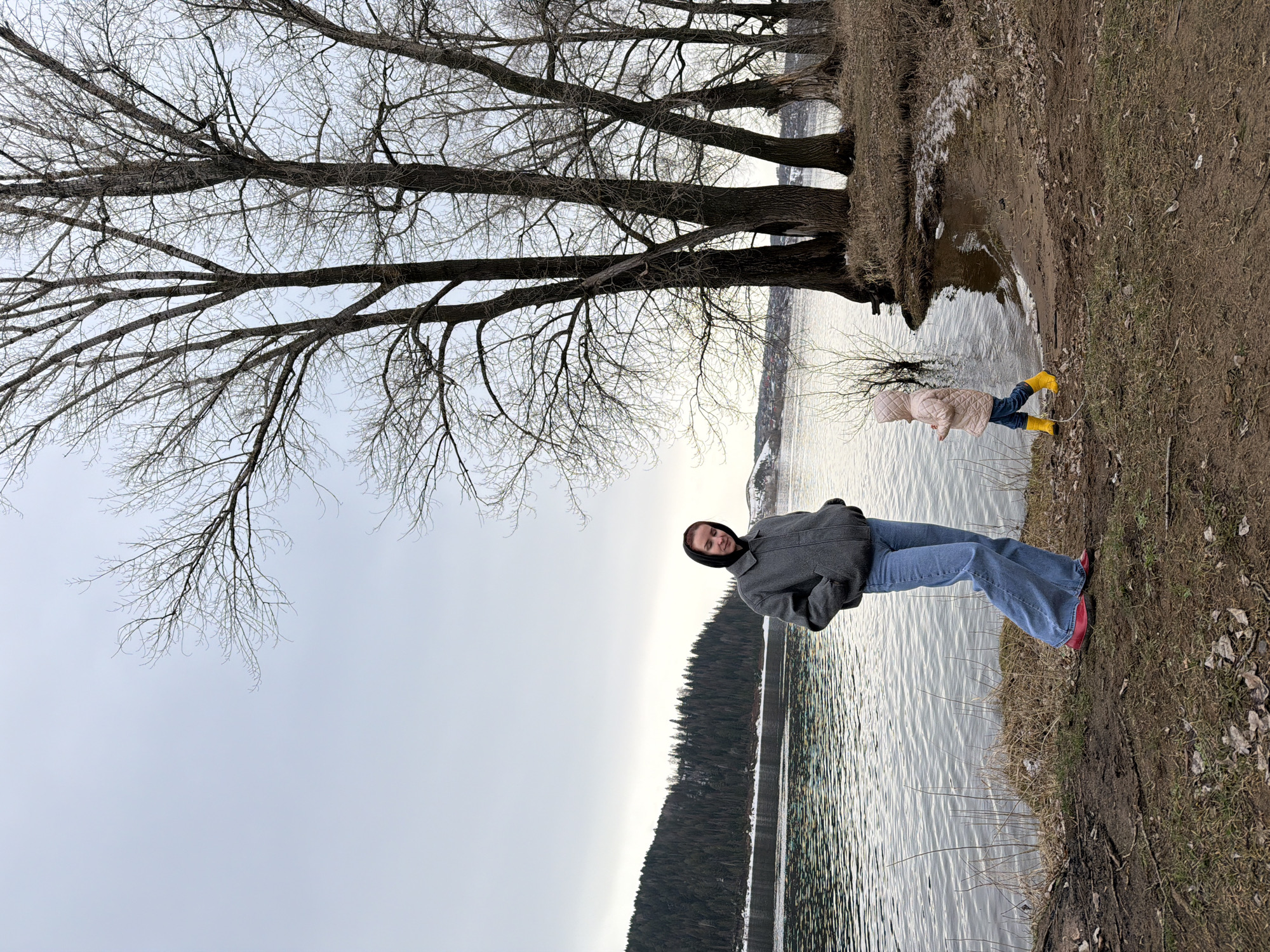 The image depicts an outdoor scene on what appears to be the shore of a body of water in Noviy, Russia. A woman wearing a gray coat, blue jeans, and red shoes stands on the shoreline. A young child in a light pink coat and yellow boots stands near the water's edge. The water is rippled, suggesting a slight breeze. Trees, without leaves, are visible. A forested area is on the far side of the water. The sky is overcast. The ground is dirt and grass.