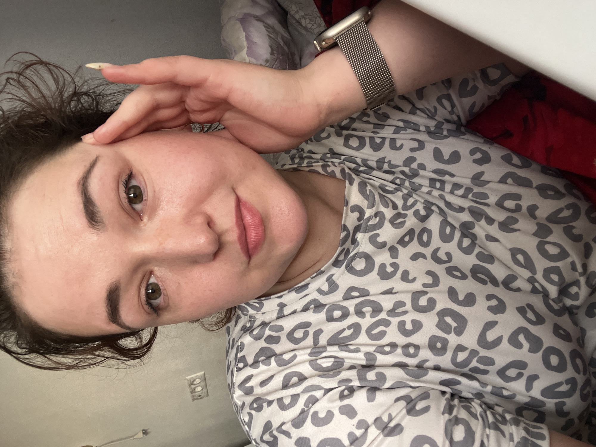 The image is a selfie taken by a young Caucasian woman with dark brown hair, green eyes, and fair skin. She is wearing a gray and white leopard print long-sleeved shirt. Her right hand is resting on her cheek and temple, with her fingers slightly extended. She is also wearing a silver mesh watch on her left wrist. The background appears to be an indoor setting, possibly a bedroom, with a wall behind her and some bedding visible. The lighting is soft and natural. There is no visible indication of the location being in Chelyabinsk, Russia.