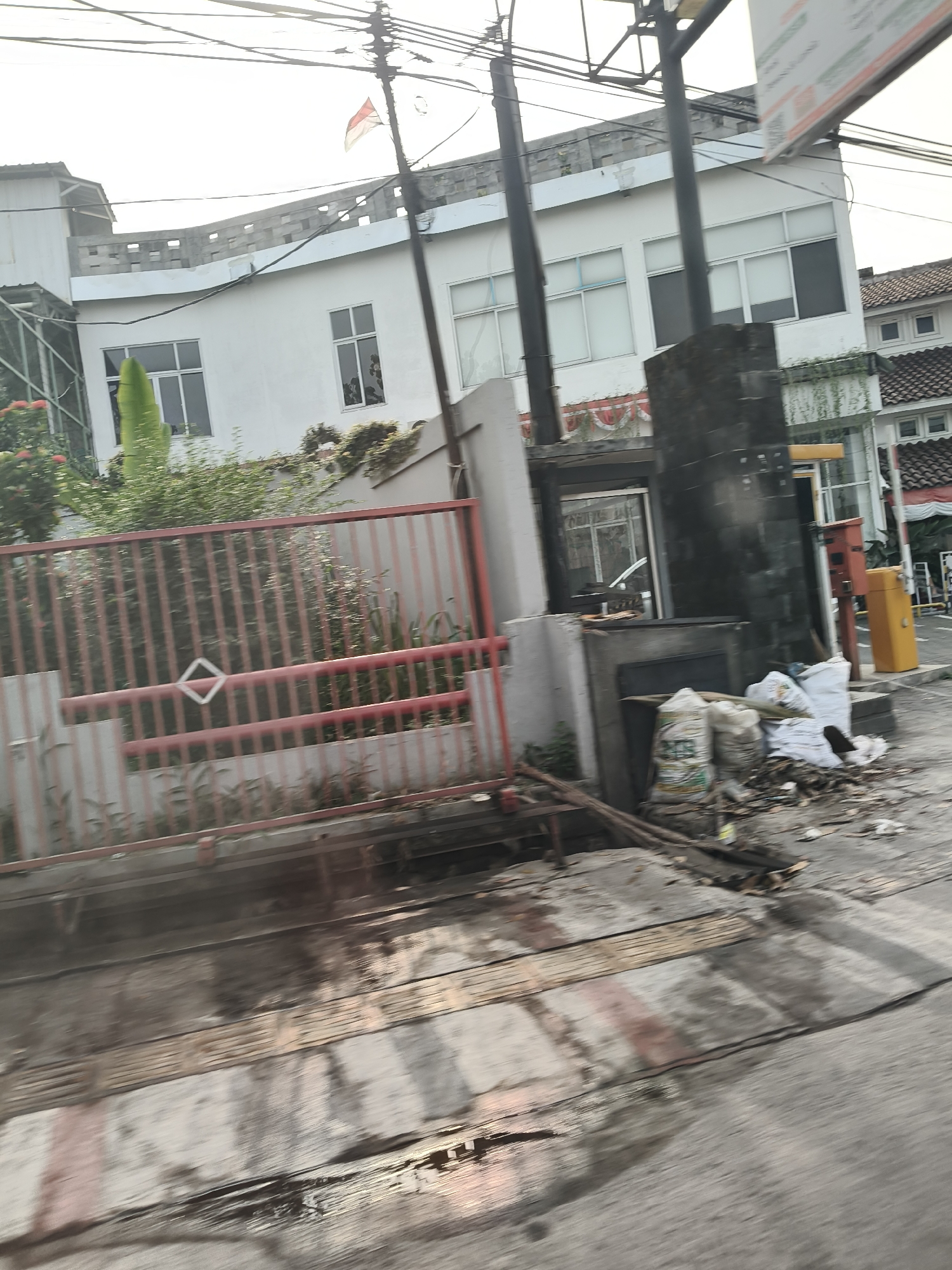 The image shows a street scene in Bandung City, Indonesia, with a white building and a red fence in the foreground. The building has several windows and a curved roofline, and a flag of Indonesia is visible on a pole. The fence is made of metal and has a diamond-shaped design. In front of the building, there are some plants and trees. The street in front of the fence is wet and appears to be made of concrete and wood. There are some sacks of material piled up near the fence. The overall impression is of a somewhat cluttered urban environment.