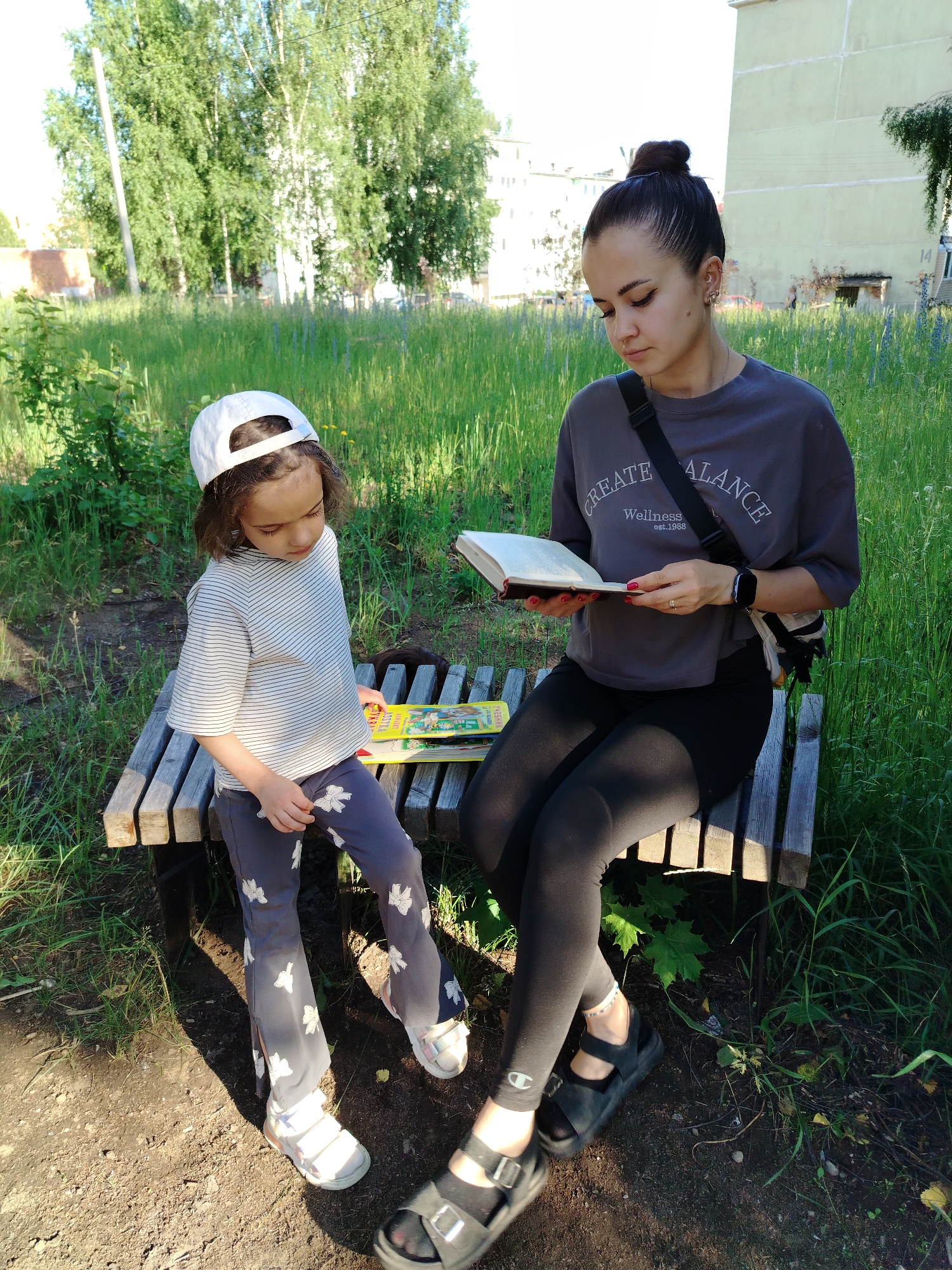 In Noviy, Russia, a young girl and a woman are sitting on a wooden bench outdoors. The girl, wearing a striped t-shirt, grey pants with a floral pattern, and a white baseball cap, is looking down at her hands. The woman, dressed in a dark grey t-shirt and black leggings, is holding an open book and reading. She has her hair pulled up in a bun. Several books are stacked on the bench between them. The background consists of tall green grass, trees, and a light-colored apartment building. The scene appears to be a park or a residential yard on a sunny day.