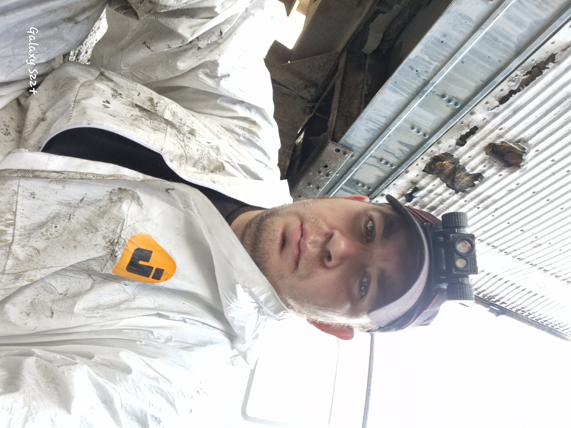 An outdoor, close-up, selfie-style photograph shows a man wearing a white disposable coverall suit, a black t-shirt, and a baseball cap. He is wearing a headlamp attached to his cap. The man's face is visible, and he appears to be looking at the camera. The coverall suit is stained with dirt and grime. The background shows a section of a corrugated metal structure that appears to be damaged and rusted. The lighting suggests it is daytime. The image is tagged "Galaxy S22+". The specific location in Zheleznogorsk-Ilimsky, Russia, is not determinable from the visual information provided in the image.