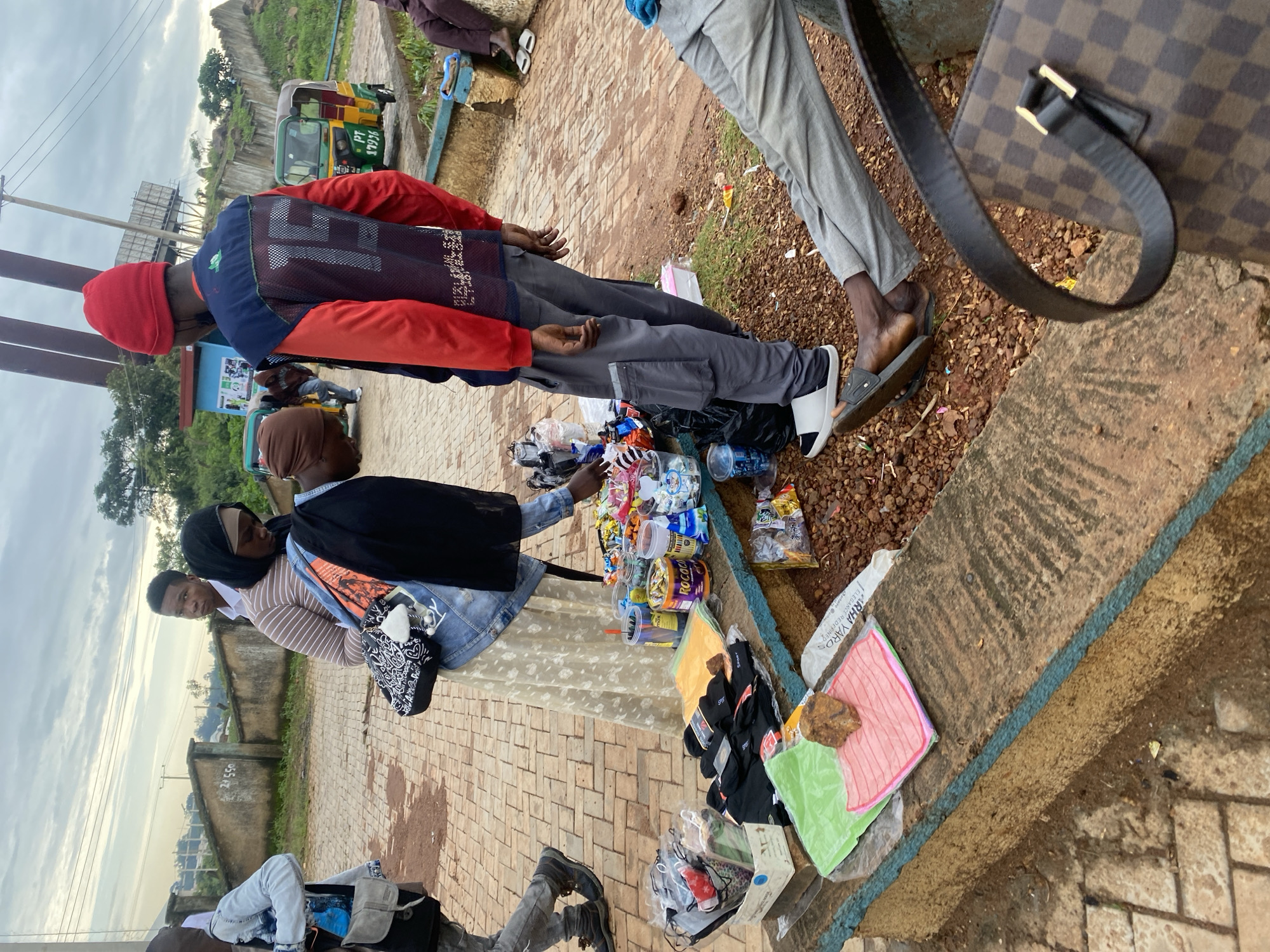 In Rafin Par, Nigeria, a street vendor is selling merchandise from a low, concrete bench. The vendor, dressed in a red beanie, red and blue jacket, and grey pants, is interacting with a customer. The customer, wearing a striped long-sleeved shirt, skirt, and denim jacket, is examining items on the bench. The bench is laden with various goods, including packaged socks, colorful cloths, canned drinks, and small bags of snacks. Behind the vendor and customer, two tuk-tuks are parked on a paved area. The environment appears to be outdoors, with a cloudy sky overhead and sparse vegetation in the background. The ground is a mix of paved surfaces and loose gravel. Other individuals are visible in the background, some appearing to be customers or bystanders. A patterned handbag is prominently displayed in the foreground.