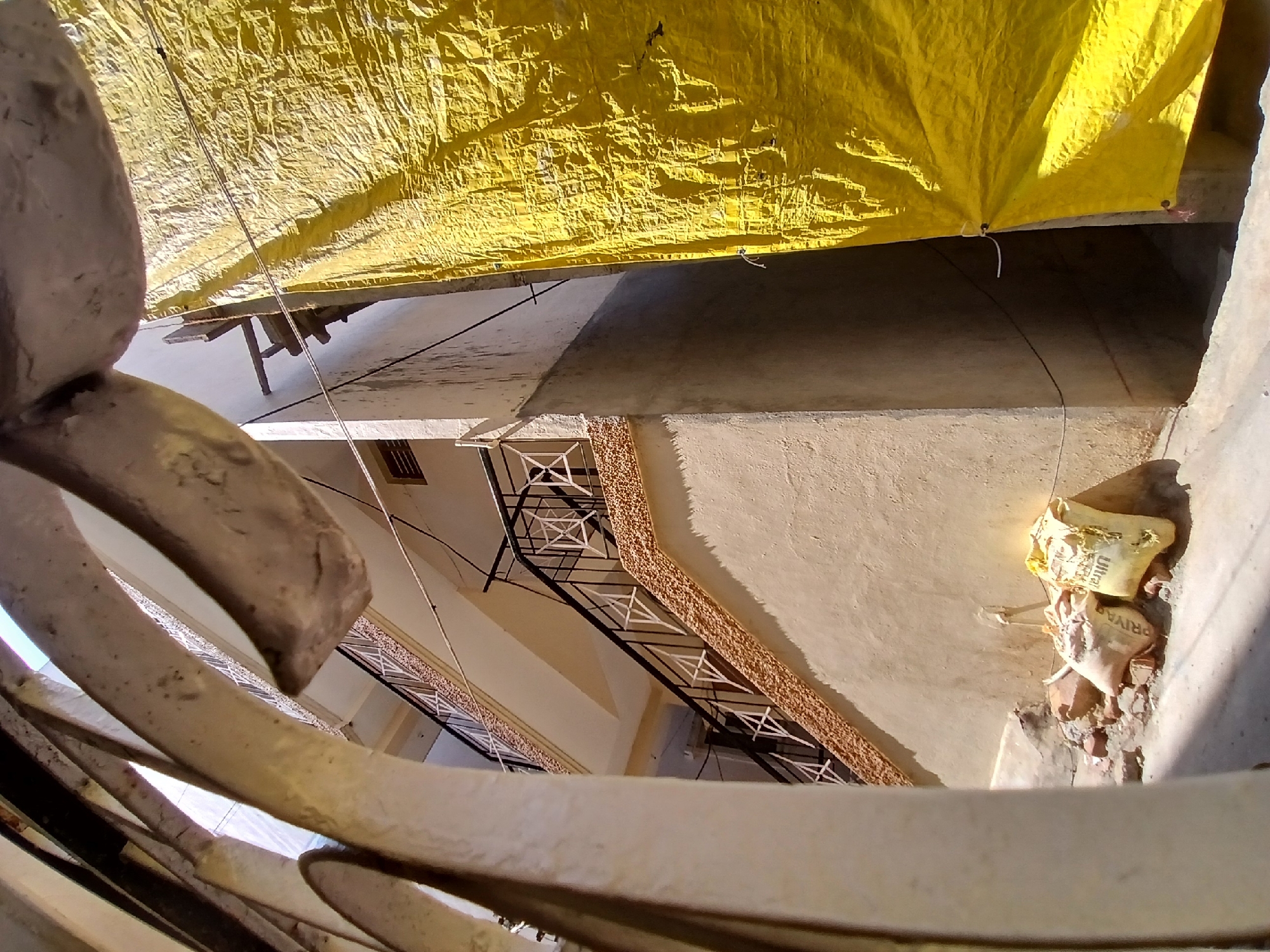 The image shows the exterior of a building in Bengaluru, India. A large yellow tarp is suspended above the courtyard. Worn cement sacks with Indian brand names like "Ultra" and "Priya" sit on a ledge. A metal railing is prominently featured in the foreground. A multi-story building's courtyard is visible, with a decorative railing, a staircase, and weathered walls. The photograph's perspective is tilted upward.