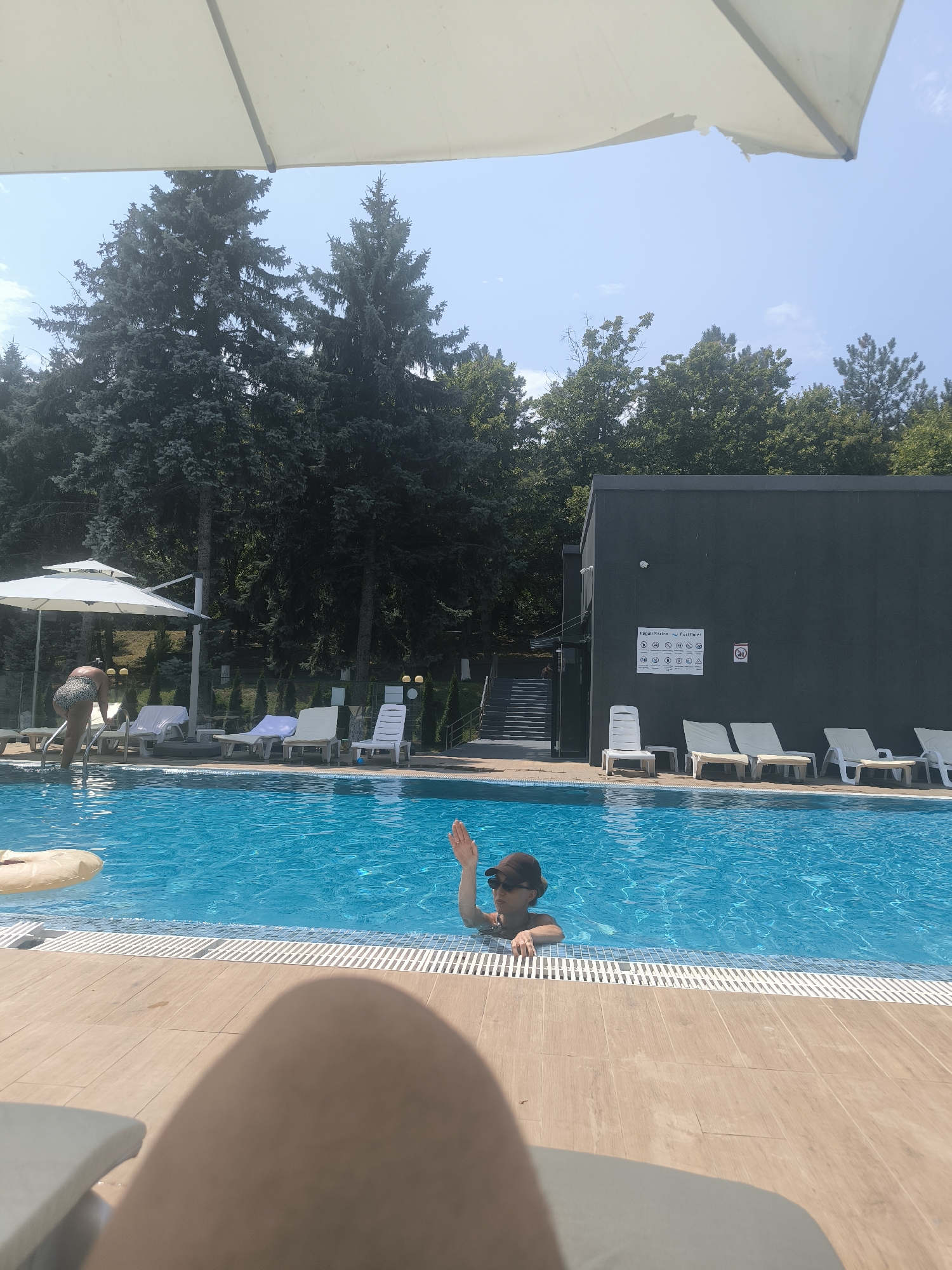 The image depicts a sunny day at an outdoor swimming pool. In the foreground, a person's leg is visible, suggesting they are lounging on a chair. The pool is filled with bright blue water. A person wearing a cap and sunglasses is in the pool, waving. To the left, a woman in a patterned swimsuit is stepping into the pool. Several white lounge chairs are arranged around the poolside. Behind the pool, there is a dark-colored building with a sign displaying various pictograms and text, likely pool rules. Tall green trees form a backdrop to the scene. The overall environment suggests a recreational area, possibly a hotel or a public pool facility. The presence of trees and the style of the building do not specifically identify the location as Ustia, Moldova, without further information.