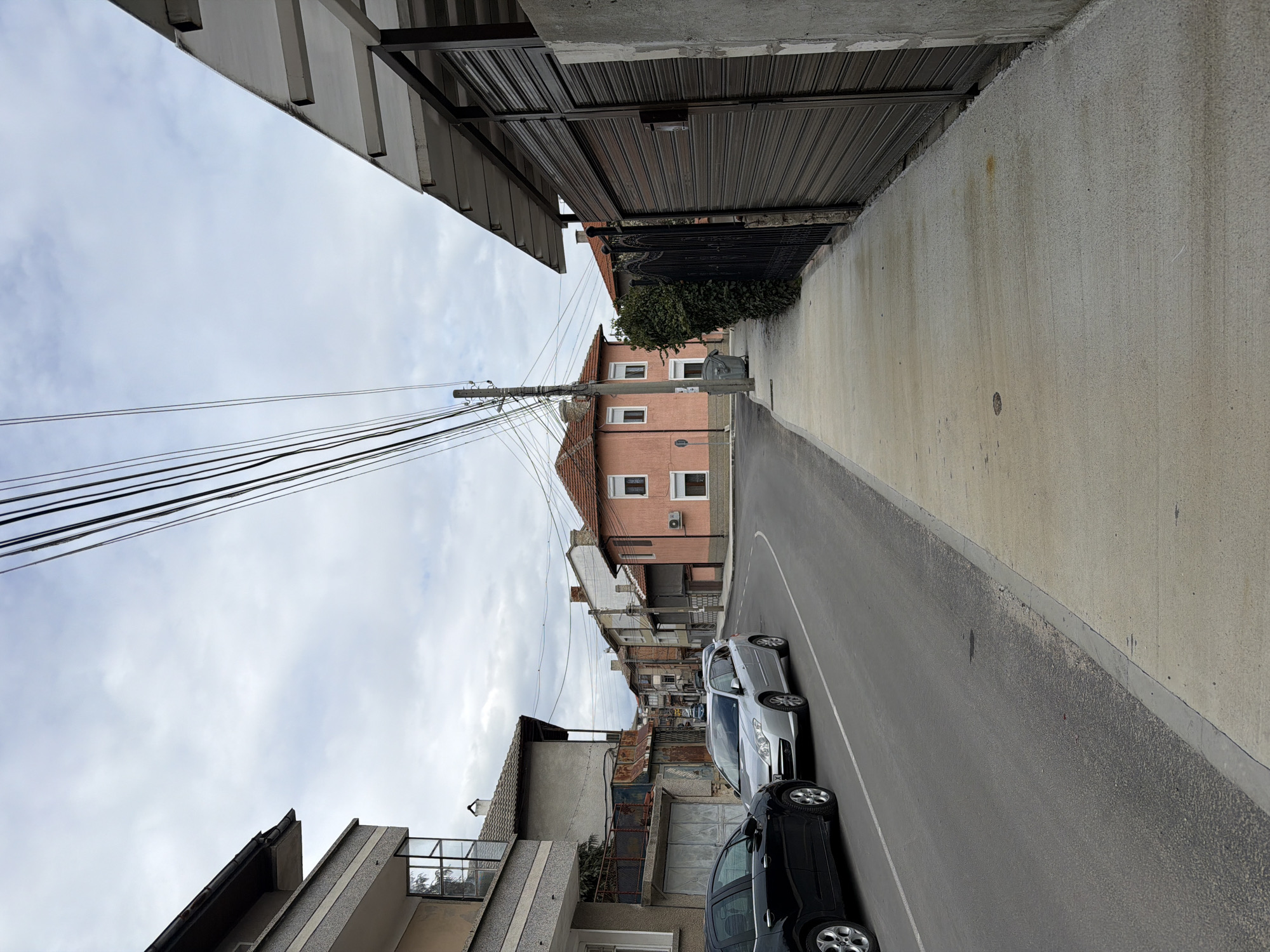 The image depicts a narrow street in Panagyurishte, Bulgaria. The view is looking down the street, with buildings lining either side. On the left is a residential building with a balcony, and on the right, there's a high concrete wall and a corrugated metal awning. Parked along the right side of the street are two cars: a silver sedan and a black SUV. Overhead, there are numerous power lines running across the street. In the distance, a pink building with a red-tiled roof can be seen. The sky is overcast. The street appears clean and well-maintained.