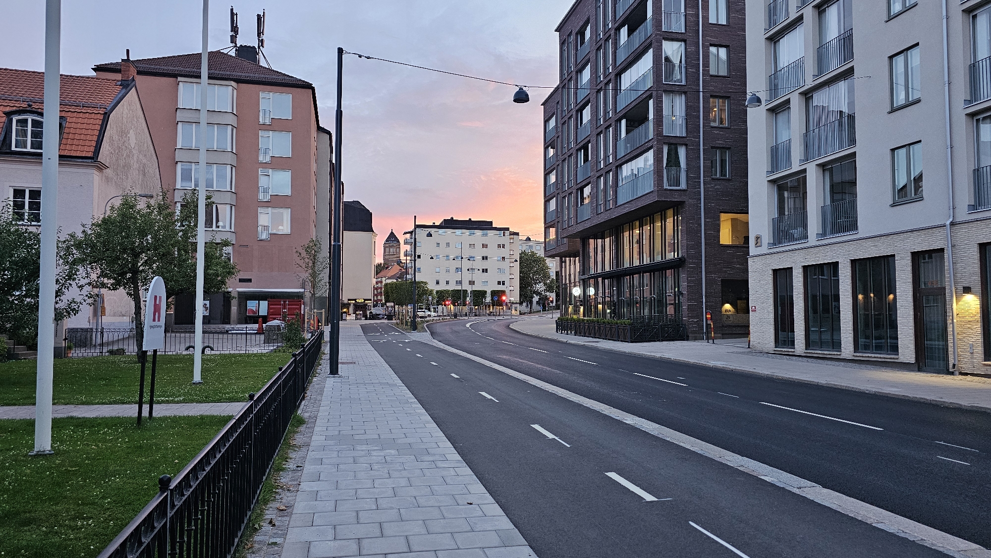 This media file captures a street scene in Linköping, Sweden, at dusk. The sky displays hues of orange and pink from the setting sun, indicating it is early evening. The weather appears to be clear.
The scene is dominated by modern and older residential buildings lining a curving street. On the left, an older, salmon-colored building with a red-tiled roof stands next to a lighter-colored building with a prominent arched window. In front of these buildings, a grassy area is bordered by a black metal fence and contains a sign with a red "H" logo and text that appears to be a business name. Several white poles, possibly flagpoles or lamp posts, are also present in this area.
To the right of the street, a contemporary dark brick building with multiple balconies and large glass windows extends along the sidewalk. Next to it is a lighter-colored building with white brick detailing on the lower level and a similar arrangement of windows and balconies above. Streetlights are visible, and the street itself is paved and marked with white lines, suggesting it is a well-maintained road. There is a bike lane marked with white dashed lines.
In the distance, more buildings are visible, including one with a distinctive tower, and traffic lights are lit. The street appears to be largely empty of people and vehicles, contributing to a quiet, serene atmosphere. The overall impression is one of urban development, blending older architecture with new construction.