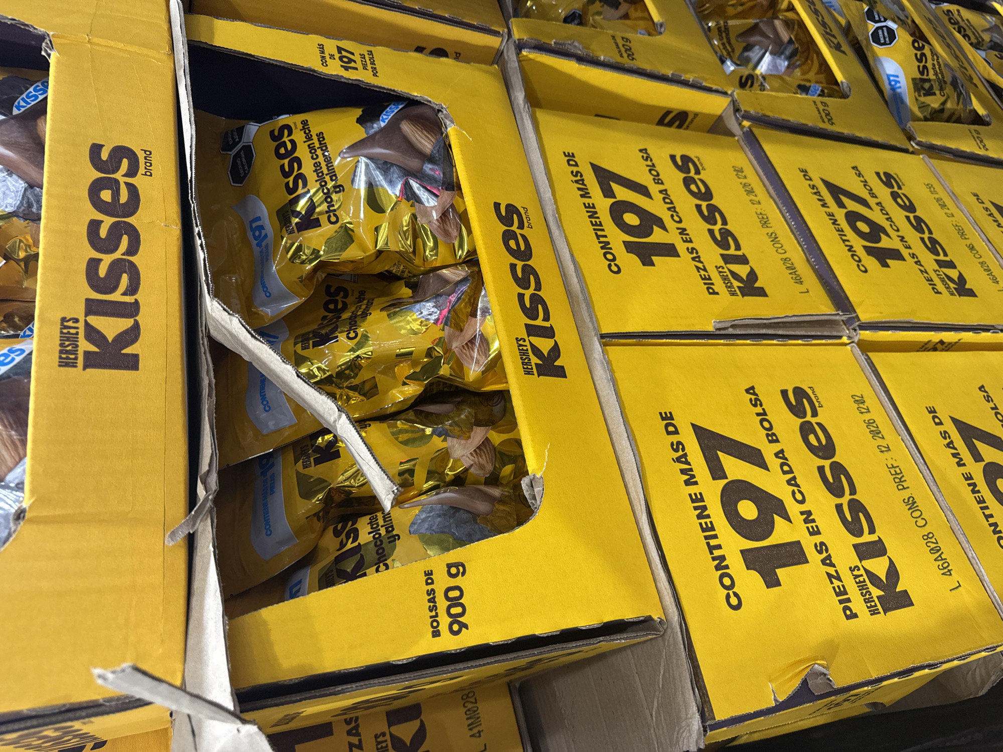 The image shows a close-up, stacked display of Hershey's Kisses chocolate bags, presumably for sale. The packaging is primarily yellow, featuring the brand name "HERSHEY'S Kisses" in black lettering and the phrase "CONTIENE MÁS DE 197 PIEZAS EN CADA BOLSA" (contains more than 197 pieces in each bag). Some bags contain milk chocolate with almonds ("Chocolate con leche y almendras"). The bags are 900 grams each. A printed expiration date, "L 46A028 CONS. PREF.: 12 2026 12:02," is visible on some of the boxes. The location and city cannot be determined from the image.