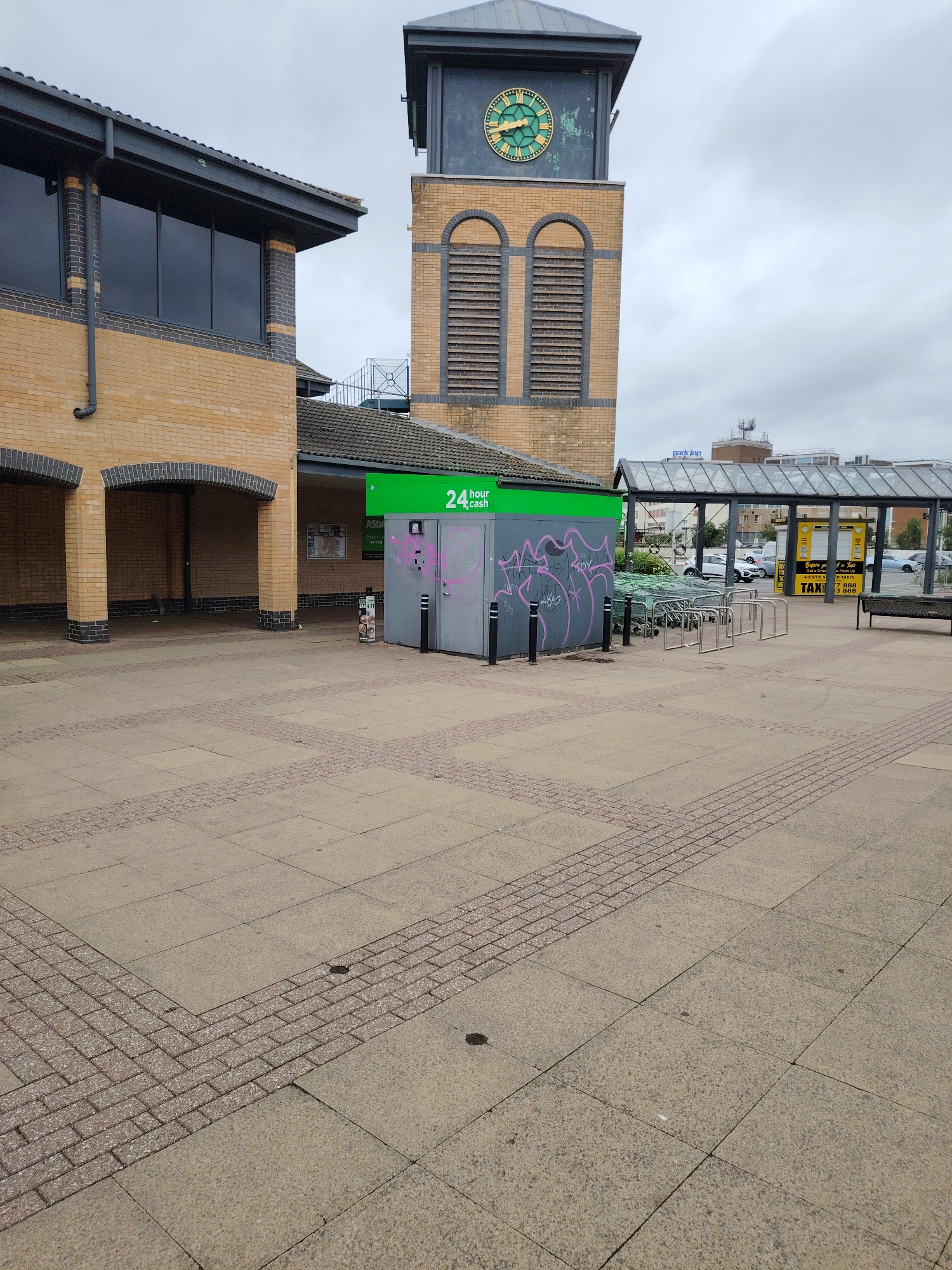 This detailed image captures an outdoor urban scene in Peterborough, United Kingdom, specifically the paved area outside the Asda supermarket on Rivergate, with the prominent Asda clock tower visible.
The foreground is dominated by a pedestrian plaza paved with light grey concrete slabs, interspersed with decorative bands of smaller reddish-brown rectangular bricks laid in a basketweave pattern. To the left, a portion of a two-story building features light tan brickwork with dark grey brick accents, dark-tinted rectangular windows on its upper level, and two arched recessed openings at ground level. A dark grey gutter and downpipe are visible on this structure.
In the mid-ground, a grey utility box, approximately 2 meters tall, stands with a vibrant green sign above it displaying "24 hour cash" in white text. The box itself is covered with irregular pink spray-painted graffiti. Three black metal bollards are positioned in front of it. Adjacent to the box, an array of silver-coloured metal bicycle racks is present, along with a stack of approximately eight green-framed supermarket trolleys. Further to the right, a contemporary shelter with a dark metal frame and glass panels covers a section of the pavement. Beyond this shelter, a yellow freestanding sign with "TAXI" and partial phone numbers is visible.
Dominating the mid-ground background is a tall clock tower constructed from light tan bricks with dark grey detailing. It features a large circular green clock face with gold hands, indicating a time between 10:20 and 10:25. Below the clock face are two tall, arched openings fitted with dark grey horizontal louvers. The top section of the tower is dark grey.
In the distant background, several vehicles are parked on a street. A multi-story building, identified by a "Park Inn" sign on its roofline, is visible against an overcast, light grey sky. No individuals are present in the scene.