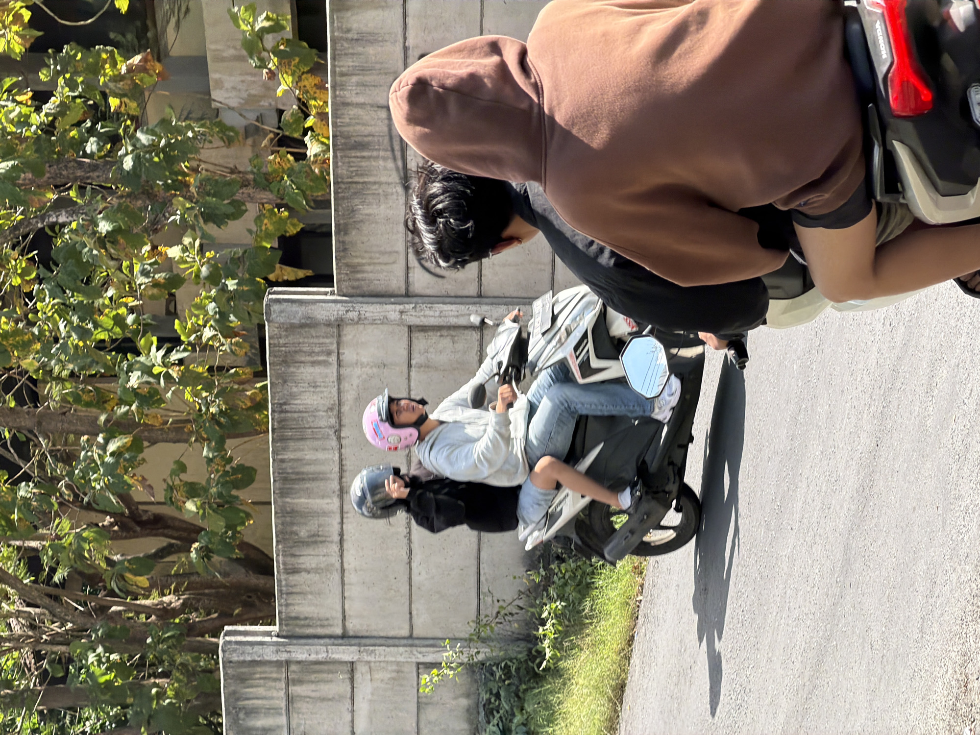 In Kuta Selatan, Indonesia, on a bright, sunny day, two individuals are riding a scooter. The person in the front, wearing a pink helmet and a light-colored hoodie, is driving. The person behind them, wearing a dark helmet and a black jacket, is a passenger. Both are looking forward as they travel down a paved road. A concrete wall, partially covered by lush green foliage, forms the backdrop. The scene is a typical street view in a tropical urban environment.