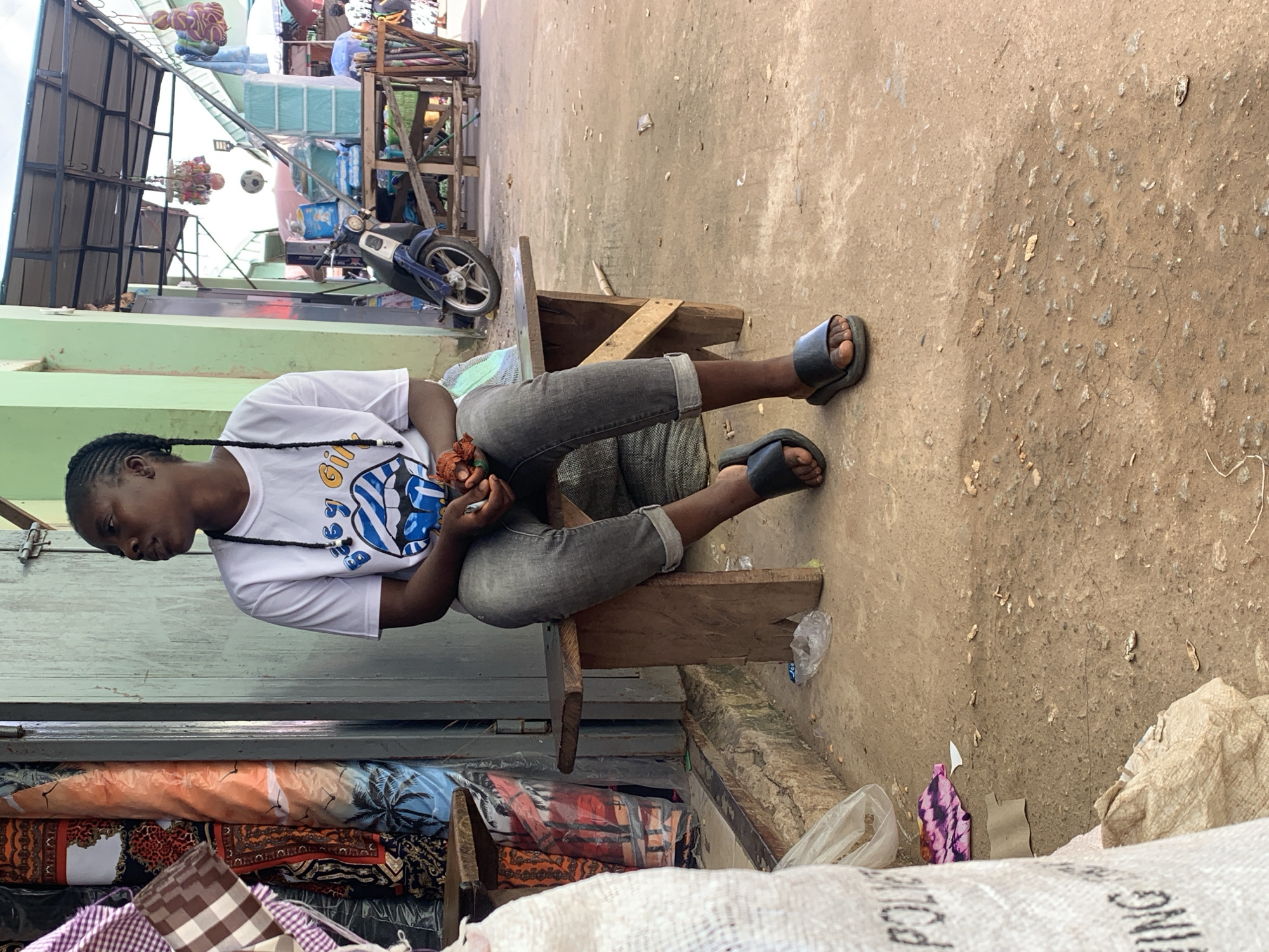 A person sits on a wooden stool on a dirt ground in what appears to be an outdoor market or street stall. The individual wears a white t-shirt with a blue and yellow graphic on it, dark grey jeans, and black flip-flops. Their hair is braided. They are holding something in their hands, possibly produce or goods for sale, and looking down at it with a neutral expression.
Behind the person, there's a stack of colorful textiles or fabrics. To the left, a green wall is visible, and above that, structures made of metal and wood, possibly part of a market stall or building. A blue and white motorcycle is parked in the background. The scene appears to be outdoors, with the ground being predominantly dirt and some rubble. The lighting suggests it is daytime. The overall atmosphere is that of a busy, perhaps informal, marketplace.